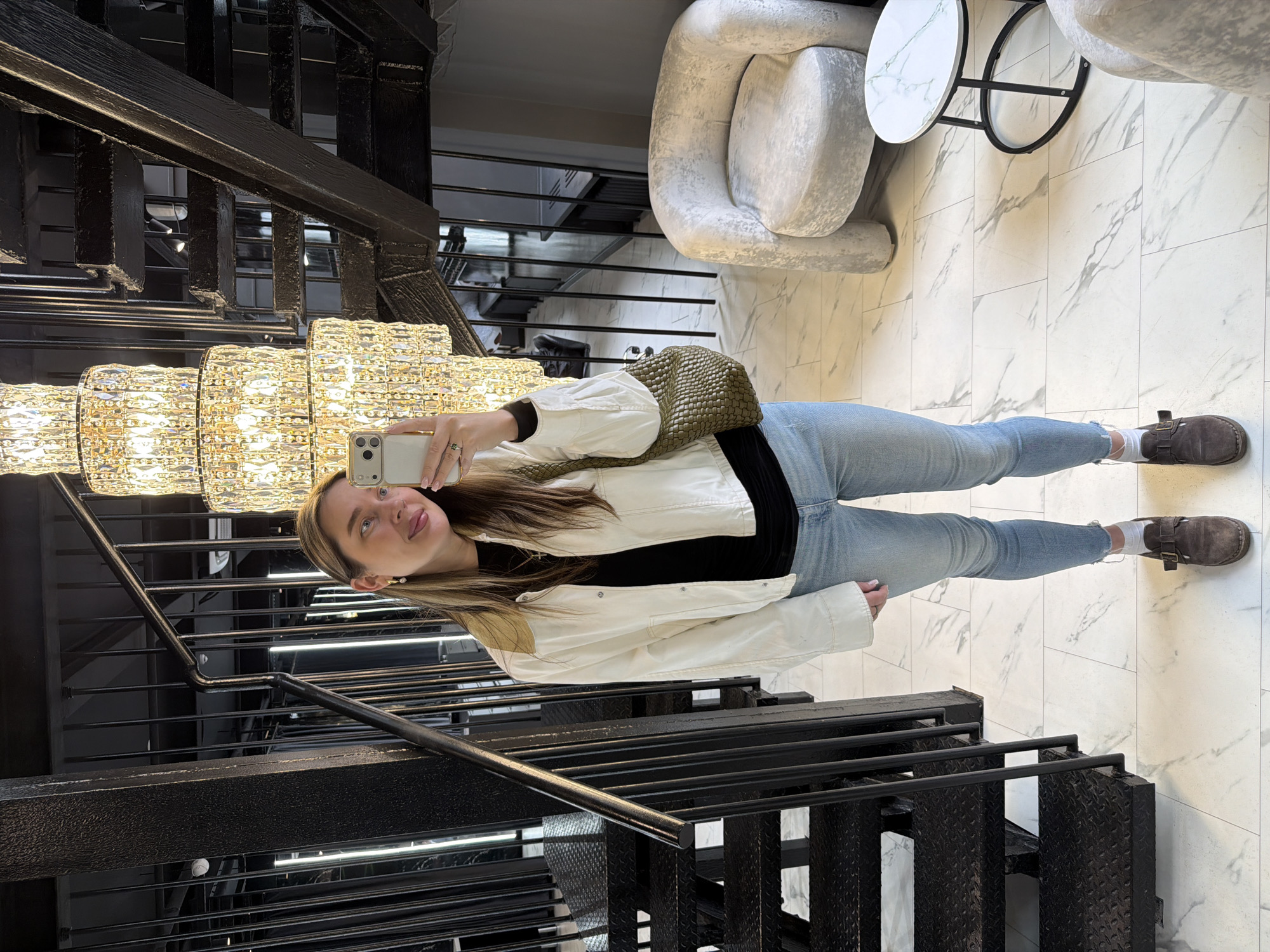 A young woman is captured in a mirror selfie, standing in a modern indoor space in Moscow, Russia. She is positioned slightly to the left of the frame, smiling with her tongue playfully extended, and holding a light-colored smartphone to take the photo. Her long, light brown hair falls over her shoulders. She wears a black top, a cream or off-white collared jacket, light blue distressed skinny jeans, and dark brown suede-like mary jane shoes with white socks. A woven dark green shoulder bag hangs on her left side. A ring is visible on her right ring finger and she has a small pearl earring.
The background features a grand, multi-tiered crystal-like chandelier emitting warm light, positioned to her left. A prominent industrial-style staircase with dark metal railings and diamond-plate treads runs along the left and bottom portions of the image. The floor is covered in large rectangular white tiles with grey marble veining. To the right of the woman, two modern, sculptural armchairs upholstered in a light grey textured fabric are visible, along with a small round side table featuring a white marble-patterned top and a black metal base. The overall aesthetic suggests a contemporary and stylish interior, possibly a lobby or upscale lounge area. No visible text is present in the image.