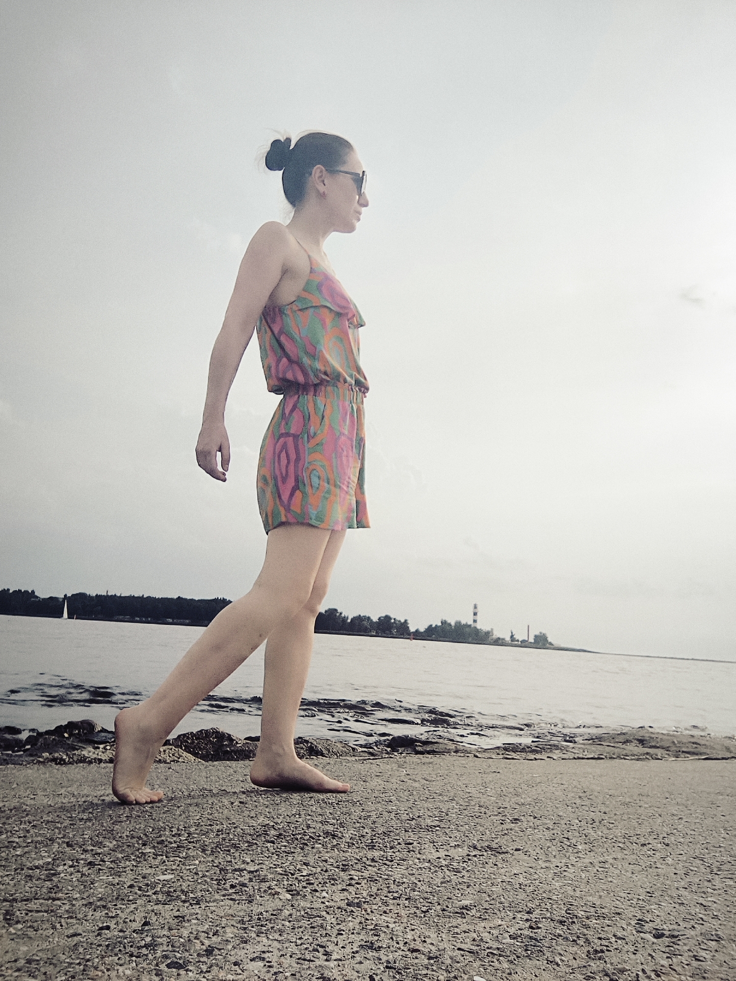 A woman with her hair in a bun and wearing sunglasses and a colorful romper stands on a gravelly shoreline, facing right. She is barefoot, with her left foot raised as if mid-stride. Behind her, the water extends to the horizon, where a dark tree line is visible, with structures that appear to be industrial or a lighthouse in the distance. The sky is overcast and bright. The scene is likely near the Daugava River in Riga, Latvia, given the presence of industrial elements and the riverine environment.