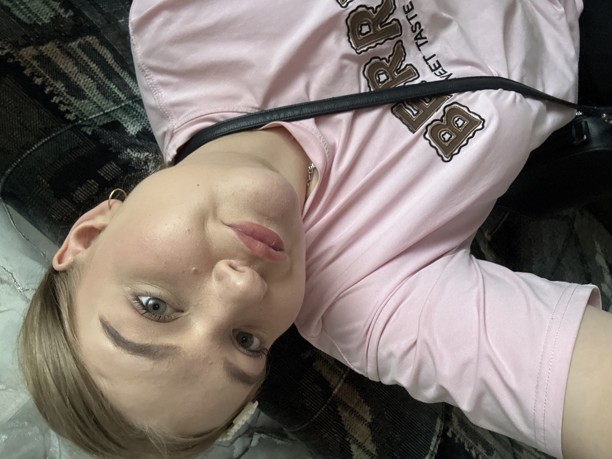 A young woman with blonde hair and light skin is lying on a patterned surface, wearing a pink t-shirt with the word "Burger" printed on it. A black strap is visible across her chest. Her face is turned towards the camera, revealing green eyes and pink lips. The background includes a patterned blanket or upholstery. There is no indication of the city of Ольховка, Russia, in the image.