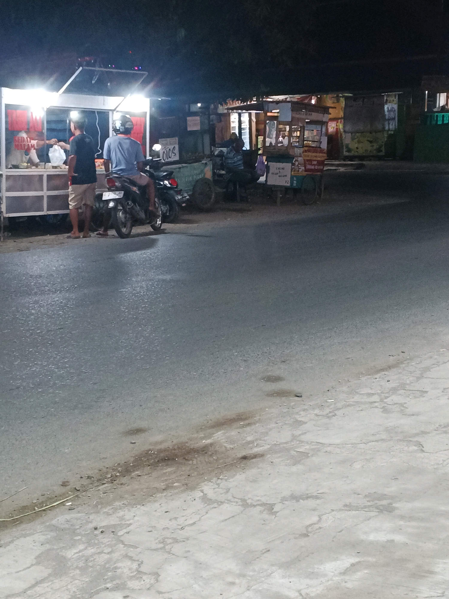 This is a nighttime photograph taken on a paved road in Taman, Indonesia, near a food stall. Two young men are present, one standing and one seated on a motorcycle. The standing man, wearing a dark t-shirt and tan shorts, faces a food stall where a person in a white shirt is handing him a white plastic bag. The man on the motorcycle, wearing a blue t-shirt and a helmet, is facing away from the camera, also near the food stall. The food stall has bright overhead lighting, with red fabric draped behind the counter. A sign with "CIOS" is visible on a nearby cart. The road appears damp, reflecting some of the light. The background shows other food carts and dimly lit buildings.