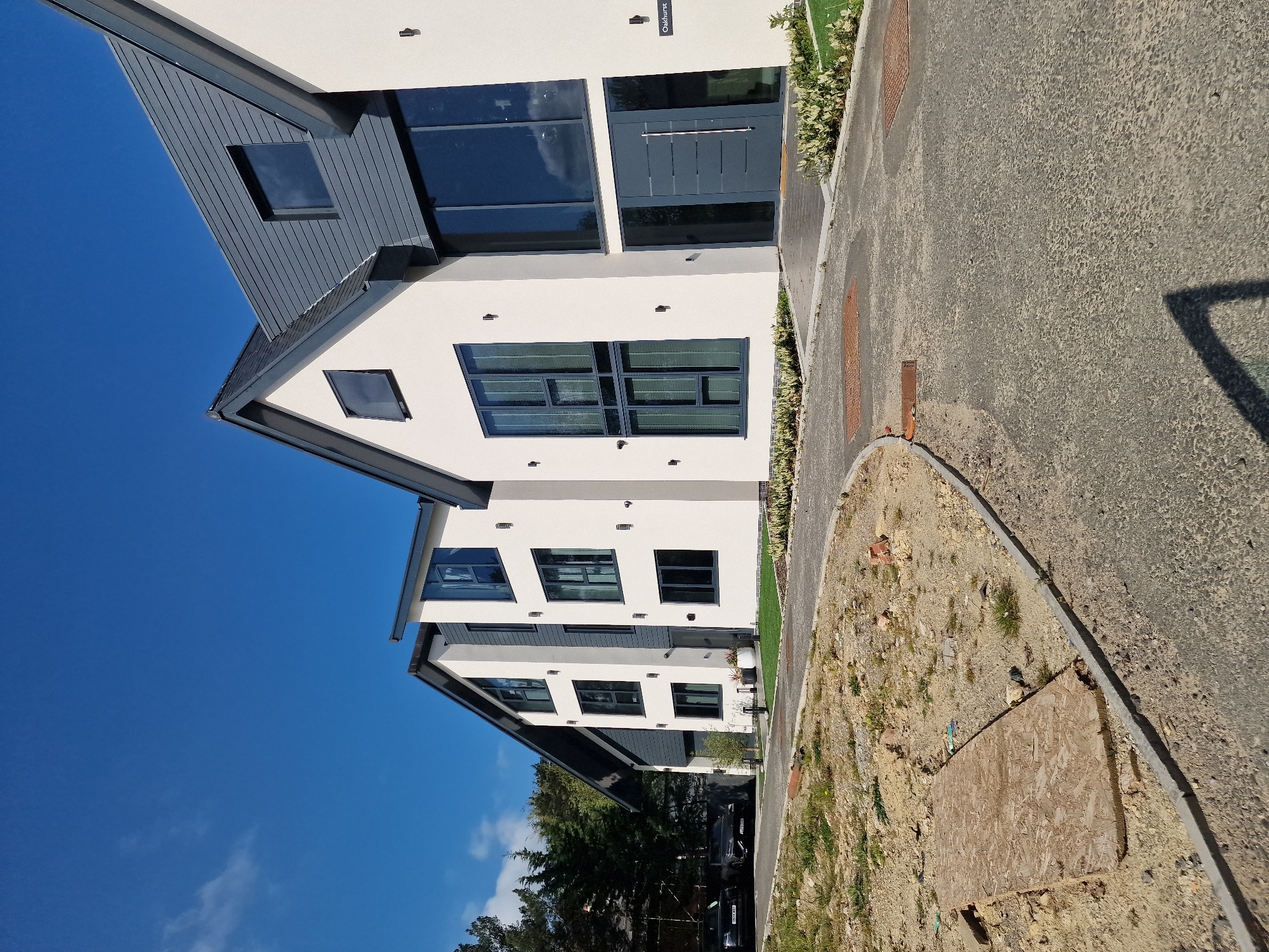 The media file displays a row of modern, newly constructed residential buildings under a bright, clear blue sky. The image is oriented sideways, with the sky occupying the upper left quadrant and the ground/path extending from the bottom right towards the top right.
The subjects are predominantly three-story houses featuring a contemporary architectural style, characterized by clean lines, white rendered facades, and dark grey window frames and cladding. Large, expansive windows, some with multiple panes, are prominent on each level. The roofs appear to be flat or very low-pitched. One house, visible in the upper mid-left, has a distinctive dark grey gable with a skylight window.
Activity is minimal, showing no people. The scene suggests a new residential development in Witton Gilbert, United Kingdom, given the fresh construction and partially unlandscaped areas.
Notable details include the bright sunlight casting sharp shadows, indicating midday or afternoon. The sky is largely cloudless, with only a few wisps of white clouds visible in the distance. The ground on the right features a paved grey path or driveway, alongside areas of bare earth with scattered small weeds and patches of grass. A wooden board lies on one such unlandscaped section. Metal grates are embedded in the paved surface. The front entrance of one house, clearly visible in a cropped hint, features a modern grey door with a long horizontal handle.
Visible text: A small black plaque above the entrance of one house reads "Oakhurst."