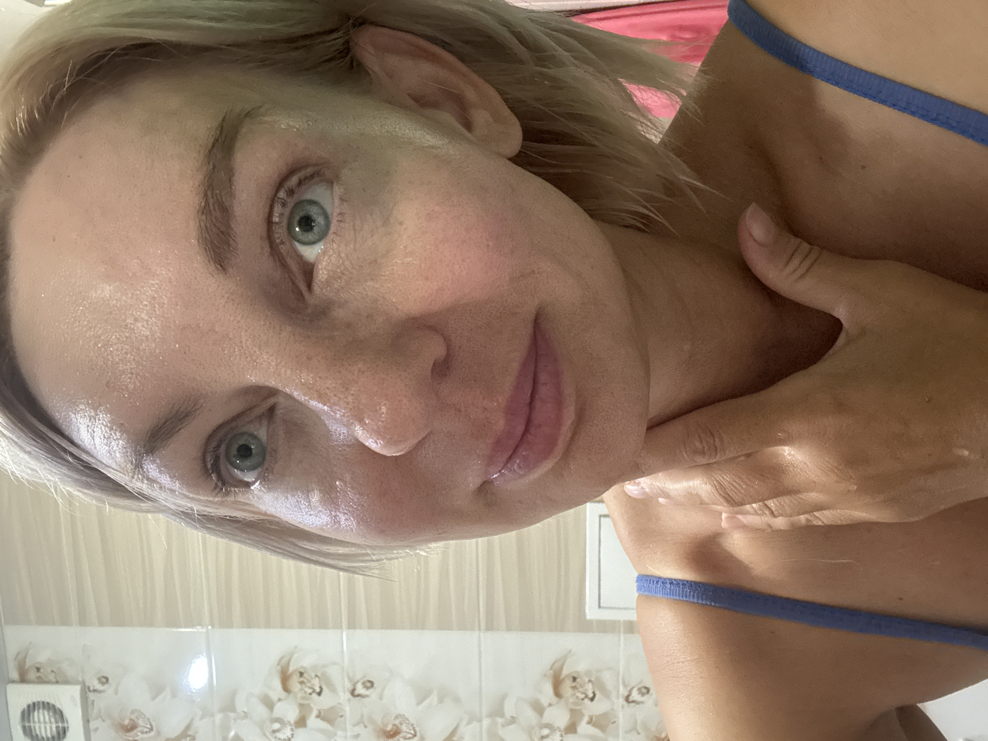 The image shows a close-up of a woman's face and upper chest. She has short, blonde hair and fair skin. Her eyes are light green and her lips are painted pink. She is wearing a blue tank top. Her right hand is touching her neck. The background appears to be a bathroom with floral patterned tiles and a white vent on the wall. The image is a selfie, likely taken in a private residence. There is no identifiable information or context provided to link this image to Sochi, Russia.