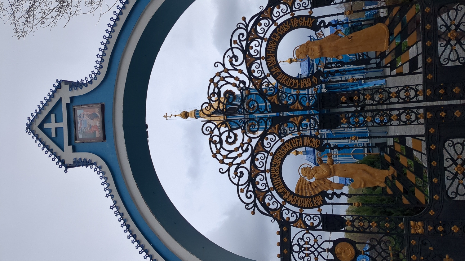 The image displays an ornate entrance gate to a religious complex, likely an Orthodox church or monastery, under an overcast sky.
The main subject is a striking blue and white archway, acting as the primary entrance. At the top of the archway, a white cross symbol is set against a blue background. Below the cross, a framed religious icon depicts multiple figures, possibly from a biblical scene. The edges of the archway are intricately decorated with a repetitive blue and white pattern.
Below the archway, a meticulously crafted black and gold wrought-iron gate is prominent. It features elaborate scrollwork and floral designs. Integrated into the gate are two golden statues, appearing to be angels or saints, each framed within a decorative circle. Around one golden figure, Cyrillic text is clearly visible, reading "БЛАГИЙ ІИСУС" (Blessed Jesus). Around the second figure, fragments of Cyrillic text can be deciphered, including "СЛАВА БОГУ ЕДИН-ТВОЕМУ" (Glory to God, the One and Only).
In the background, behind the gate, a vibrant blue church building with white trim and characteristic golden onion domes and finials is visible. Bare tree branches are silhouetted against the cloudy sky in the upper left portion of the image. The scene is illuminated by diffused daylight, indicative of the overcast weather. The architectural style, religious iconography, and visible Cyrillic text are strong cues for an Orthodox Christian site, consistent with the provided location context of Ulyanovsk, Russia. No specific activity or event is taking place; the image focuses on the architectural and religious elements of the entrance.