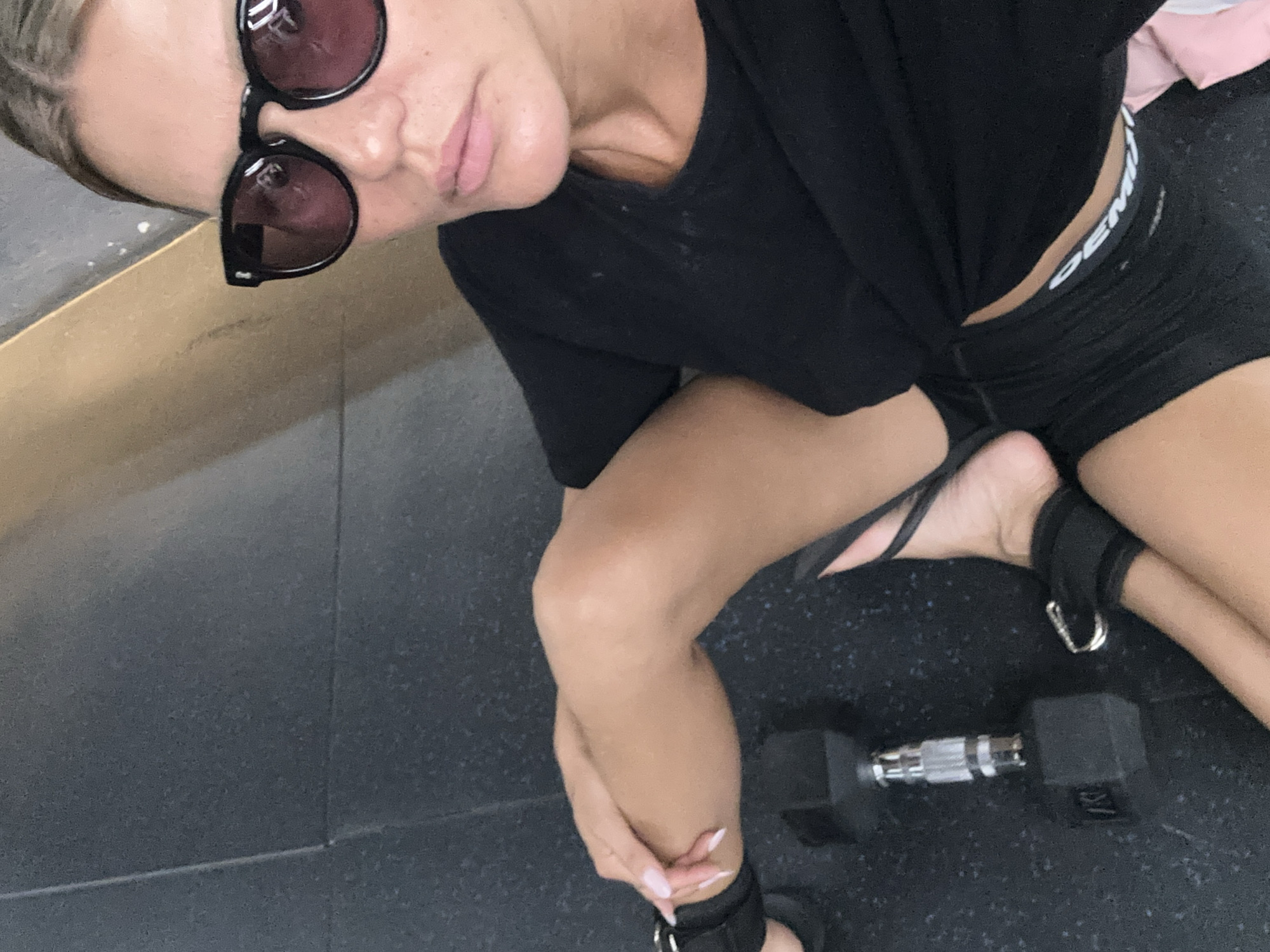 A woman is sitting on a dark gray gym floor, holding onto her leg. She is wearing a black sports bra and black shorts. She has on sunglasses. A dumbbell is on the floor next to her. The setting appears to be a gym.