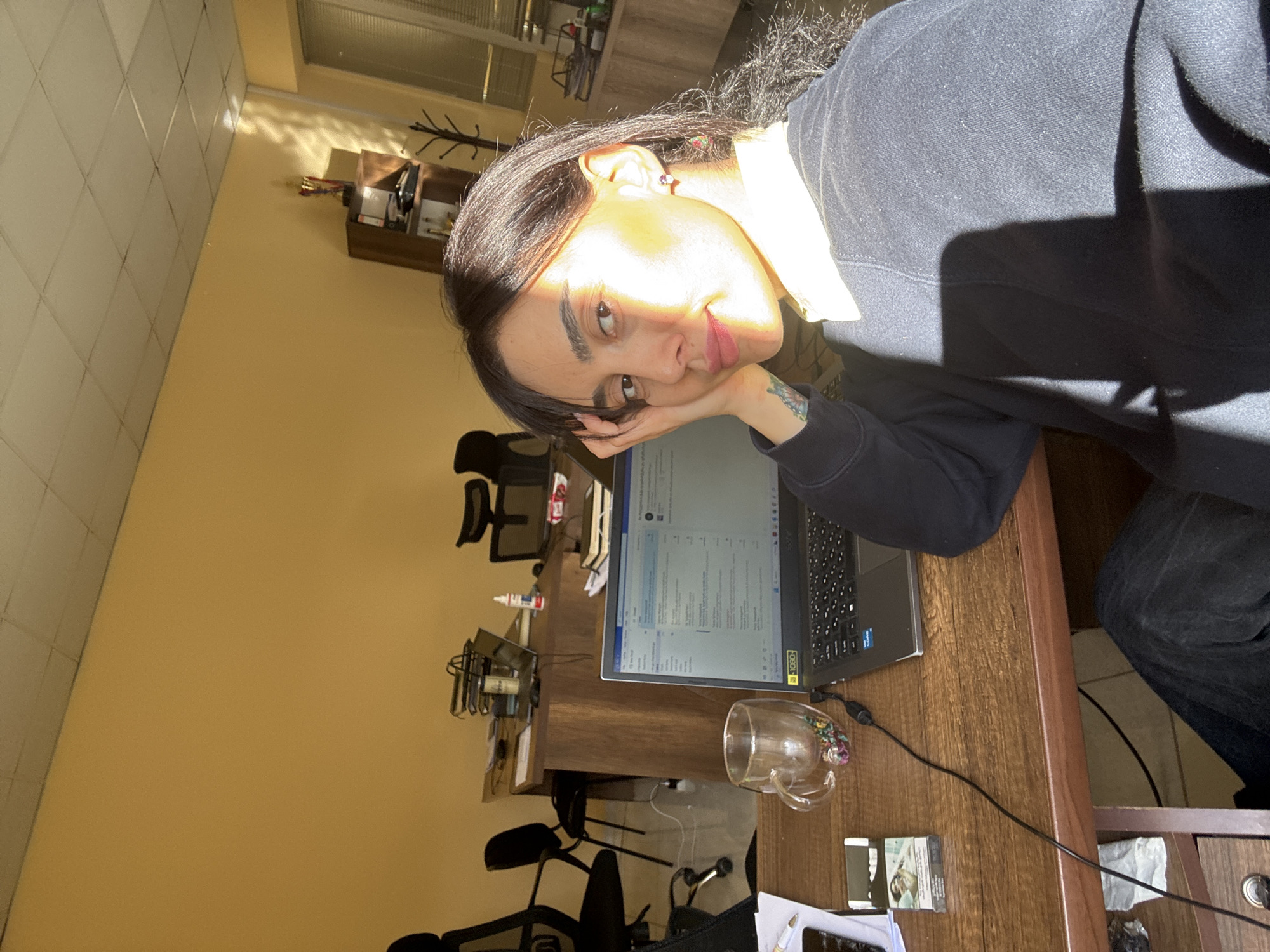 A person with dark hair and light skin is sitting at a desk with a laptop in an office setting. The person is wearing a dark sweater with a white collar peeking through and has a tattoo visible on their forearm. They are resting their face on their hand, looking at the camera. The laptop screen displays a document with lines of text. A clear glass mug sits on the desk next to the laptop. In the background, there is a yellow wall, office chairs, and shelves with various items. Based on the style of furniture and architecture, it is plausible this photograph was taken in an office in the city of Tbilisi, Georgia.