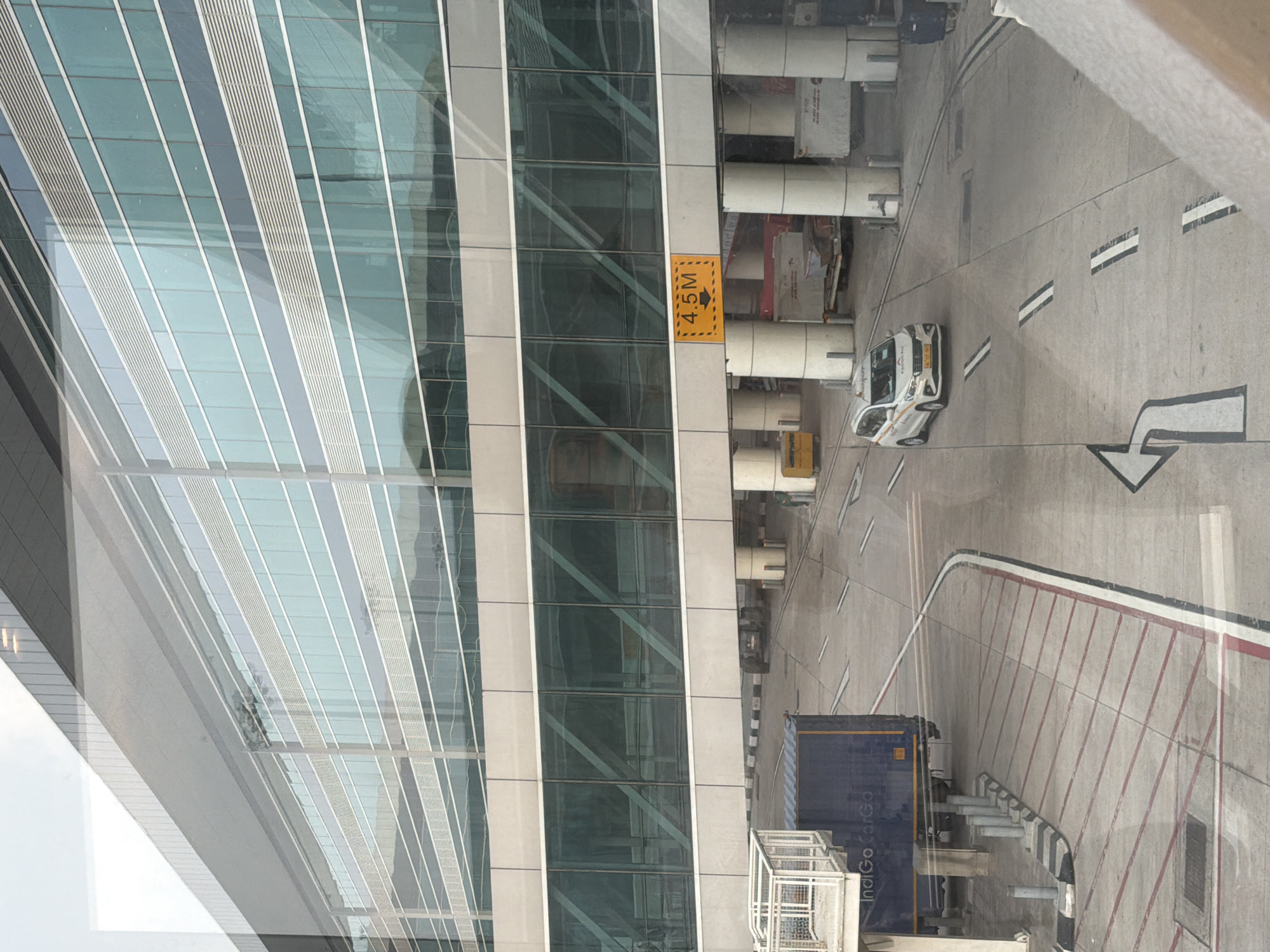 The image shows a view of a modern airport terminal building with a skywalk, taken from a window. The building features a glass facade with horizontal blue and silver stripes. Below the skywalk, there is a roadway with markings. A white taxi with the license plate "DL 7C T 6049" is driving on the roadway. In the background, there are several luggage carts and industrial-style containers. To the left, a blue cargo container with "IndiGo Cargo" printed on it is visible. A yellow sign with black text and an arrow indicating "4.5M" is mounted on a support beam of the skywalk. The scene is well-lit, suggesting daylight conditions. The location is identified as New Delhi, India.