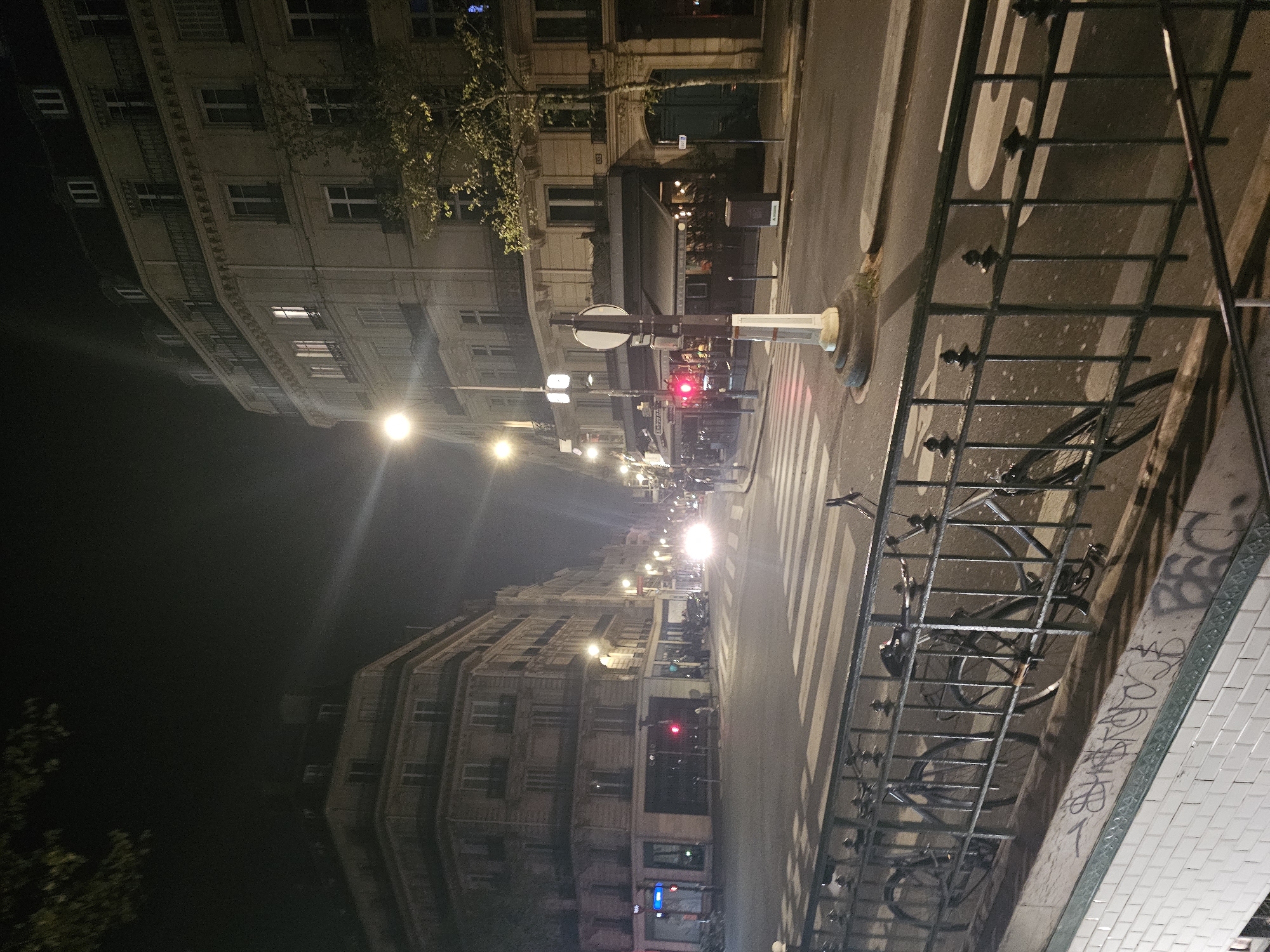 The image captures a street scene in Paris, France, at night. The view is from a slightly elevated perspective, looking down a street lined with Haussmannian buildings, characterized by their uniform facades and rows of windows. Streetlights illuminate the scene, and traffic lights are visible further down the road. A pedestrian crossing stretches across the street. On the right side of the frame, there is a wrought-iron fence with bicycles locked to it. Graffiti marks the wall next to the fence. The scene appears to be quiet and urban.