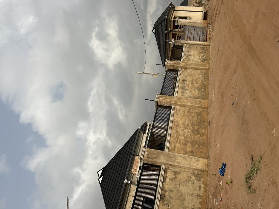 The scene depicts an exterior view in Ilishan, Nigeria, under an overcast sky. In the midground, a textured, light brown concrete perimeter wall with prominent vertical pillars runs horizontally. This wall is surmounted by a dark metal fence composed of horizontal slats, fitted with electric fence insulators and wires.
Behind the wall to the left, a portion of a two-story building is visible. This structure features a dark, possibly charcoal-gray, tiled hip roof with white trim, and a light-colored upper facade with dark horizontal window grates. An area of exposed red brickwork is also present on its facade. To the right of this, also situated behind the wall, stands a smaller, single-story, light-colored octagonal building with a matching dark tiled hip roof. Its facade incorporates a gray metal entrance gate with horizontal slats and a visible window opening to the right of the gate. A slender wooden utility pole with an overhead electrical wire is positioned behind the wall, slightly right of center.
The foreground consists of reddish-brown dirt soil with scattered patches of sparse green vegetation. A blue and white patterned fabric item, likely an article of clothing, and fragments of white and red plastic debris are visible on the dirt surface in the lower left portion of the image. The upper portion of the image is dominated by a sky heavily covered with dense gray and white clouds, with a small area of lighter blue visible through a gap in the upper-left. No individuals are present in the scene.