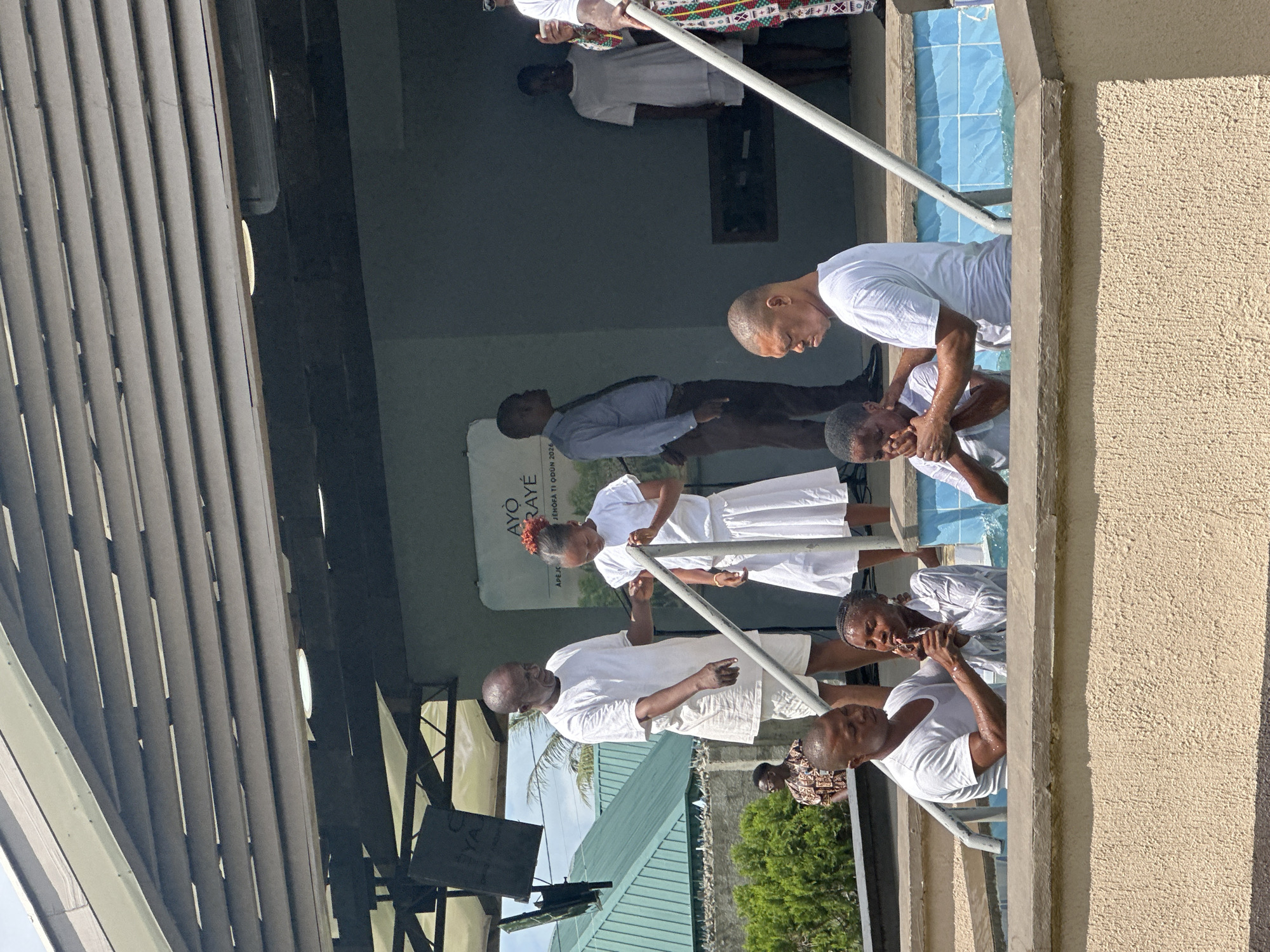 In Itoga, Nigeria, a baptismal ceremony is taking place in an outdoor setting under a covered structure. The water in the baptismal pool is turquoise and appears to be about waist-deep for those in it. Several individuals are involved in the ceremony, all dressed in white. In the foreground, a man is baptizing a woman in the pool, with his hands on her head. To the left of this pair, another woman is also being baptized by a man. Further back, a man in a white t-shirt and shorts, presumably a religious leader, is standing next to a young girl in a white dress, holding her hand as she enters the pool. Behind them, other figures are visible, including a man in a blue shirt and brown pants, and a woman in a white t-shirt and shorts. A large sign with text is visible on the wall in the background, and part of a building with green roofing is seen to the left. The overall atmosphere suggests a religious gathering focused on the sacrament of baptism.