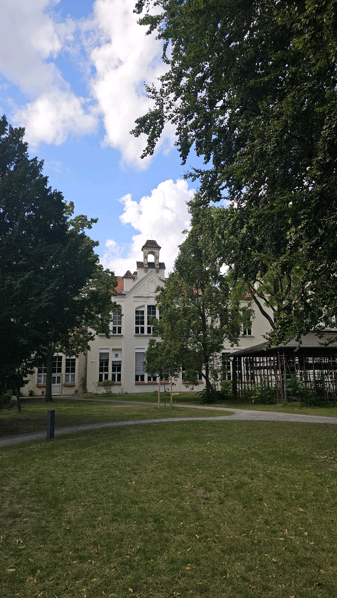 The image depicts the exterior of a white, multi-story building with a red-tiled roof, framed by lush green trees. The building features numerous large windows and a distinctive tower-like structure with an arched opening at the top. A well-maintained lawn stretches out in front of the building, with a winding gravel path. In the foreground, the grass is interspersed with fallen leaves, and a dark, upright post is visible on the left. To the right, a covered structure with wooden supports and fencing can be seen. The sky above is a vibrant blue with scattered white clouds. This building is identified as the Helmholtz-Gymnasium in Berlin, Germany. No people or actions are visible in the image.