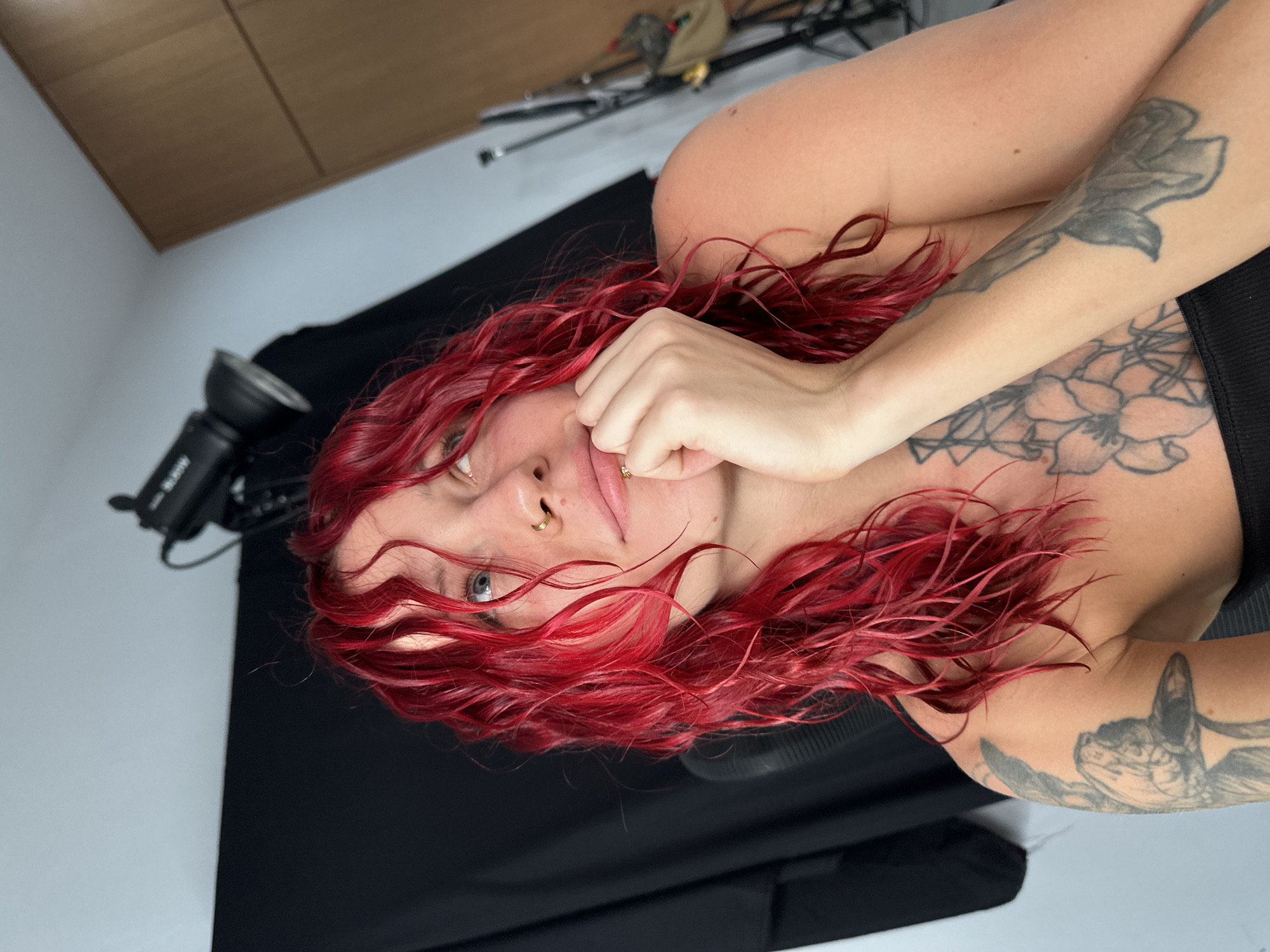 A waist-up shot of a woman with red hair and tattoos on her arms is lying on her side against a black surface, possibly a backdrop, in a room that appears to be a photography studio. A professional camera flash is partially visible in the lower left corner. A cabinet is on the top left wall, while a tripod and a bag are visible on the top of the photo. The location cannot be determined to be in Dalung, Indonesia.
