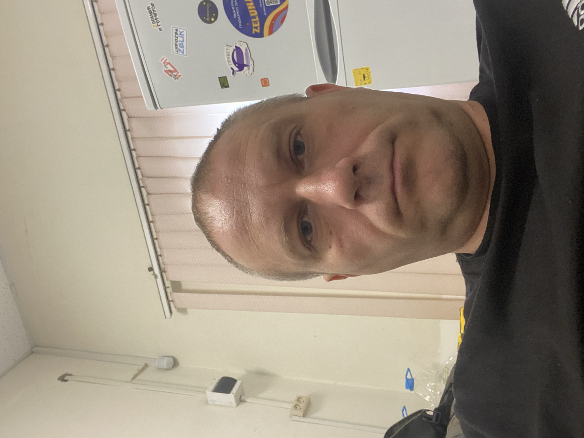 The image is a close-up, slightly angled selfie of a Caucasian man with short hair and stubble. He is wearing a black t-shirt. The man's face is the primary focus, with his expression appearing neutral or slightly amused. Behind him, a white appliance, likely a refrigerator, is visible, adorned with several stickers. One sticker features a whale and the word "ПРИВЕТ!" (Hello!). Another sticker has the text "ZELUNAR". A yellow sticker is also visible. The background appears to be an indoor setting, possibly a room or kitchen, with beige blinds and a white wall. Electrical outlets are visible on the wall. The lighting suggests it is daytime. The location is identified as Elino, Russia.