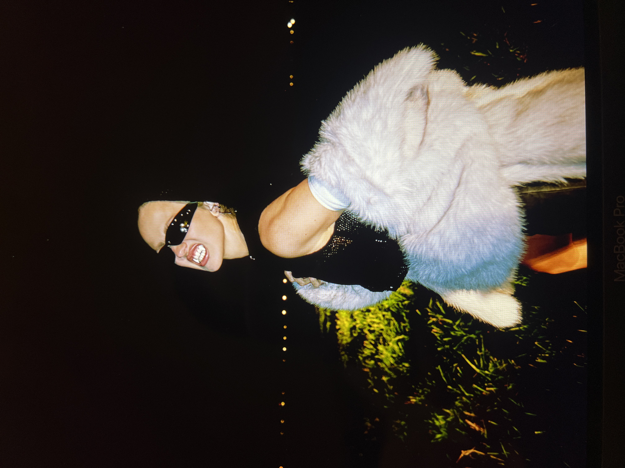 The image shows a person wearing sunglasses and a fluffy white coat at night. The person is outdoors, with some grass and lights visible in the background. The scene appears to be a still from a video or a photograph taken indoors, possibly on a laptop screen, given the presence of the "MacBook Pro" text at the bottom of the image. There is no indication of the location being Saint Petersburg, Russia.