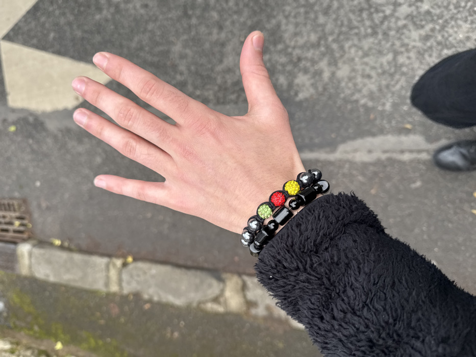 A close-up, first-person perspective image captures a fair-skinned human left hand, palm partially visible, extended forward outdoors in Orsay, France. Three beaded bracelets adorn the wrist: the top and bottom bracelets are composed of dark, spherical beads, while the middle bracelet features three prominent, round, gem-encrusted beads colored green (top), red (middle), and yellow (bottom), mimicking a traffic light sequence. The wearer's arm is covered by a black, textured, possibly fleece-like sleeve. The background consists of a damp, dark paved surface, likely asphalt, with a concrete curb and a metal drainage grate visible in the upper left quadrant. Faint lighter geometric patterns on the pavement are discernible in the upper right. A black shoe, consistent with a boot, belonging to the individual, is partially visible in the lower right corner.