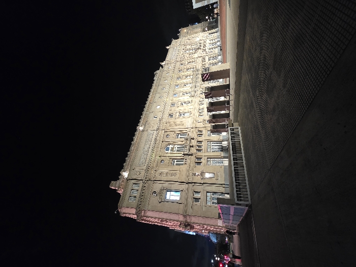 An ornate building, lit up against a night sky, dominates the left side of the frame. The building appears to be several stories tall with intricate detailing on its facade, and illuminated windows. To the right is a paved surface. At the bottom of the image, near the building, are hints of traffic lights and the presence of a road. The overall mood is dark, with the building as the primary source of light.