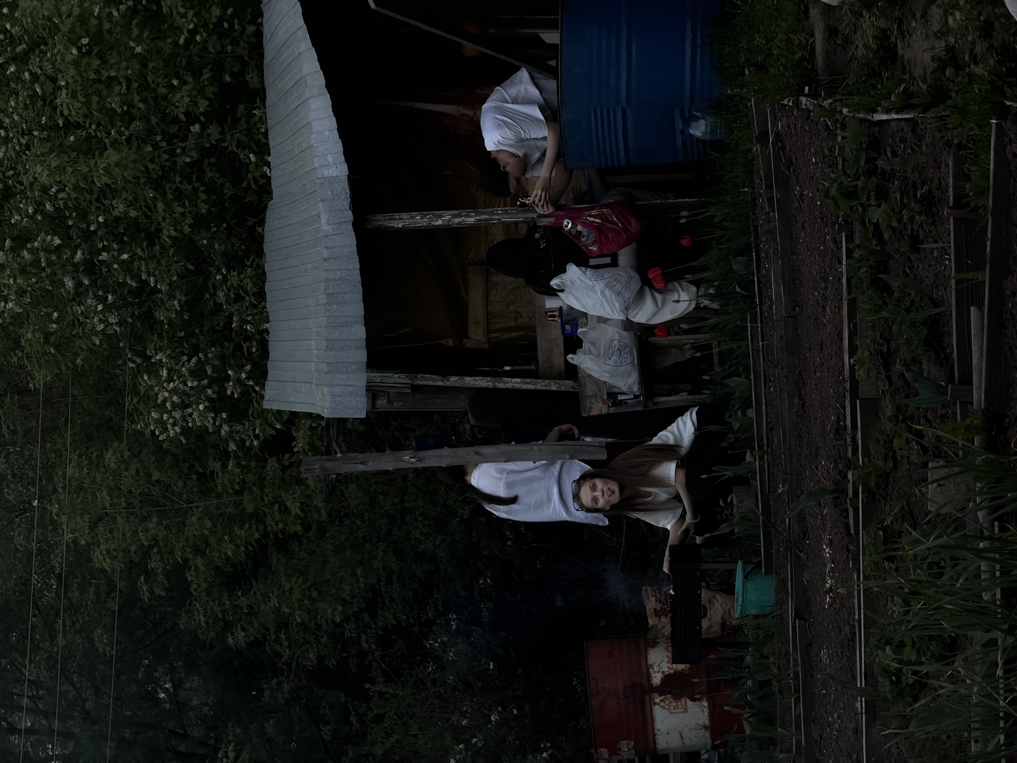 The image shows an outdoor scene in Perm, Russia. Two individuals are present: One is located inside a metal structure with a corrugated roof, appearing to use a cell phone. The other person is positioned outside and to the lower right, wearing glasses and a white top and black pants, and looking at the camera. Near that person is a barbecue or fire pit. There's abundant foliage nearby. A garden plot or raised bed extends across the right side of the frame.