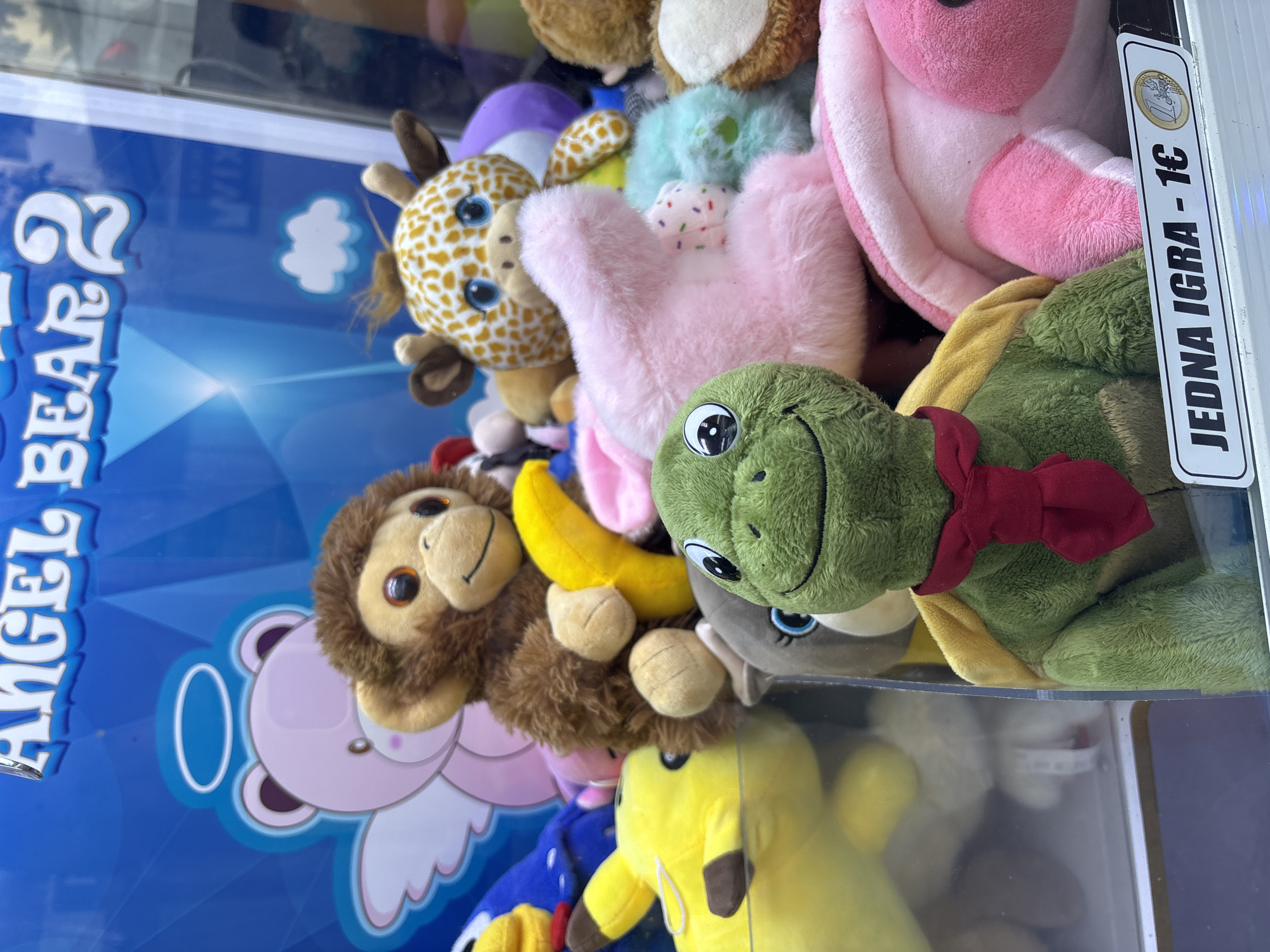 The image shows a close-up view of a claw machine filled with plush toys. The toys include a green turtle with a red scarf, a brown monkey holding a yellow banana, a giraffe, a pink bunny, a yellow Pikachu, and other various stuffed animals. In the background, a blue banner reads "ANGEL BEAR 2". A price tag at the bottom of the machine states "JEDNA IGRA - 1€" which translates to "ONE GAME - 1€". The scene is indoors, likely in a public space given the presence of an arcade game. The lighting is bright, illuminating the colorful toys. There are no people visible in the image. The location is identified as Petrovac centar, Montenegro.