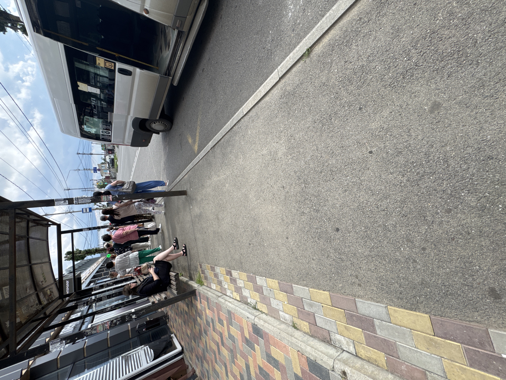 A white minibus is parked at a bus stop on a sunny day. Several people are standing in line, waiting to board the bus, while one person is sitting on a bench, looking at their phone. The bus stop has a metal frame and a transparent roof. The ground is paved with colorful interlocking tiles and asphalt. The scene appears to be in Mikhaylovsk, Russia, given the text "Михайловск" visible on a sign near the bus stop. The sky is partly cloudy, suggesting a warm day. The overall atmosphere is that of a typical urban scene, with people going about their daily routines.