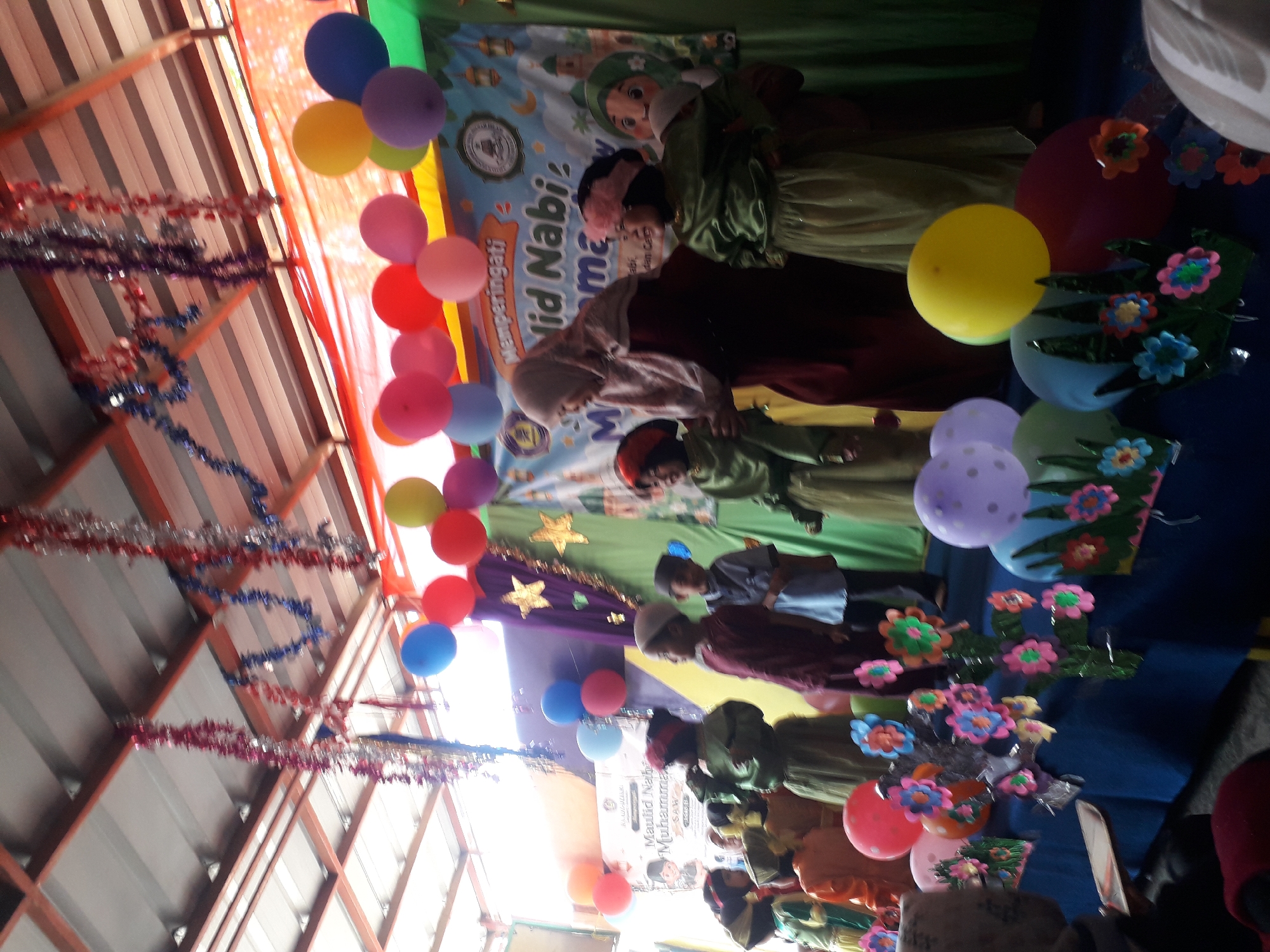 The image shows a group of children dressed in traditional clothing, likely on a stage, participating in an event. The backdrop features a banner with text that appears to be in Arabic, suggesting a religious or cultural celebration. Balloons and decorative garlands hang from the ceiling, indicating a festive atmosphere. In the foreground, several children are visible, some standing and others seated. The overall setting suggests an indoor event, possibly a school or community gathering in Cankuang, Indonesia, given the context of the prompt. The lighting is bright, and the image is taken from a slightly low angle, looking up towards the stage.