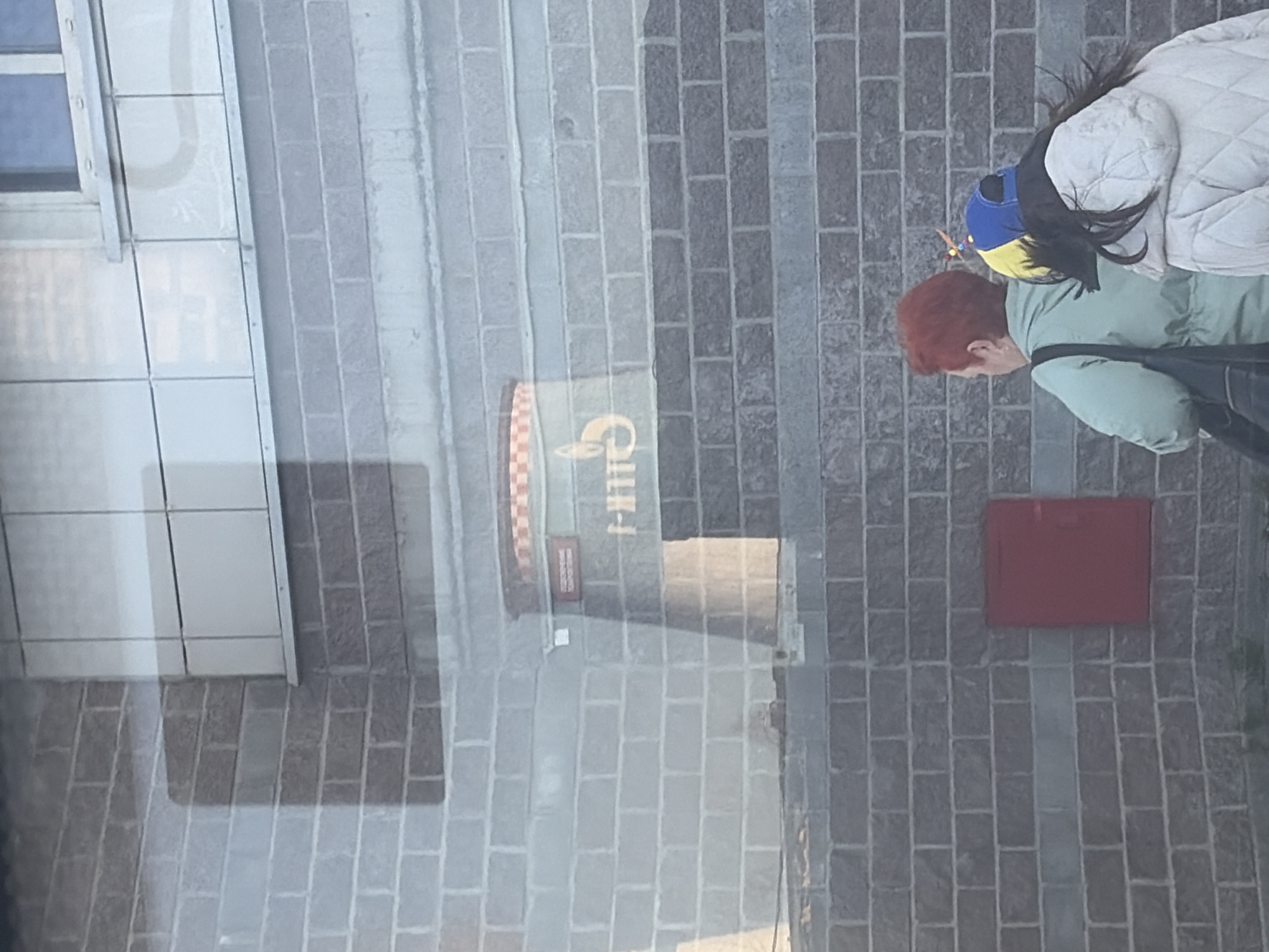 The image, rotated 90 degrees clockwise, captures an urban street scene through what appears to be a large glass reflection.
On the right side of the frame, two individuals are visible. The person in the foreground has red hair, is wearing a light green jacket, and is looking downwards. They carry a dark bag over their shoulder. Behind them, a second individual is seen from the back, wearing a cream-colored quilted jacket and a distinctive blue and yellow knitted hat.
The left side of the image features a vertical section of a modern building facade with a window, which is likely part of the reflection on the glass. The central area displays a large reflected sign with a checkered border. The mirrored Cyrillic text "СНИКЕРС" (SNICKERS) is clearly visible on this sign. Below it, smaller, illegible characters can be seen.
The background and foreground elements consist of a grey brick pattern, which due to the image's rotation, appears as a horizontal brick wall. On this brick surface, to the right of the reflected sign, a rectangular red object (possibly a mailbox or utility box) is mounted, with some faint, unreadable text on its surface. The overall lighting is soft and diffused, characteristic of an overcast day. The setting suggests a city street in Saint Petersburg, Russia.
