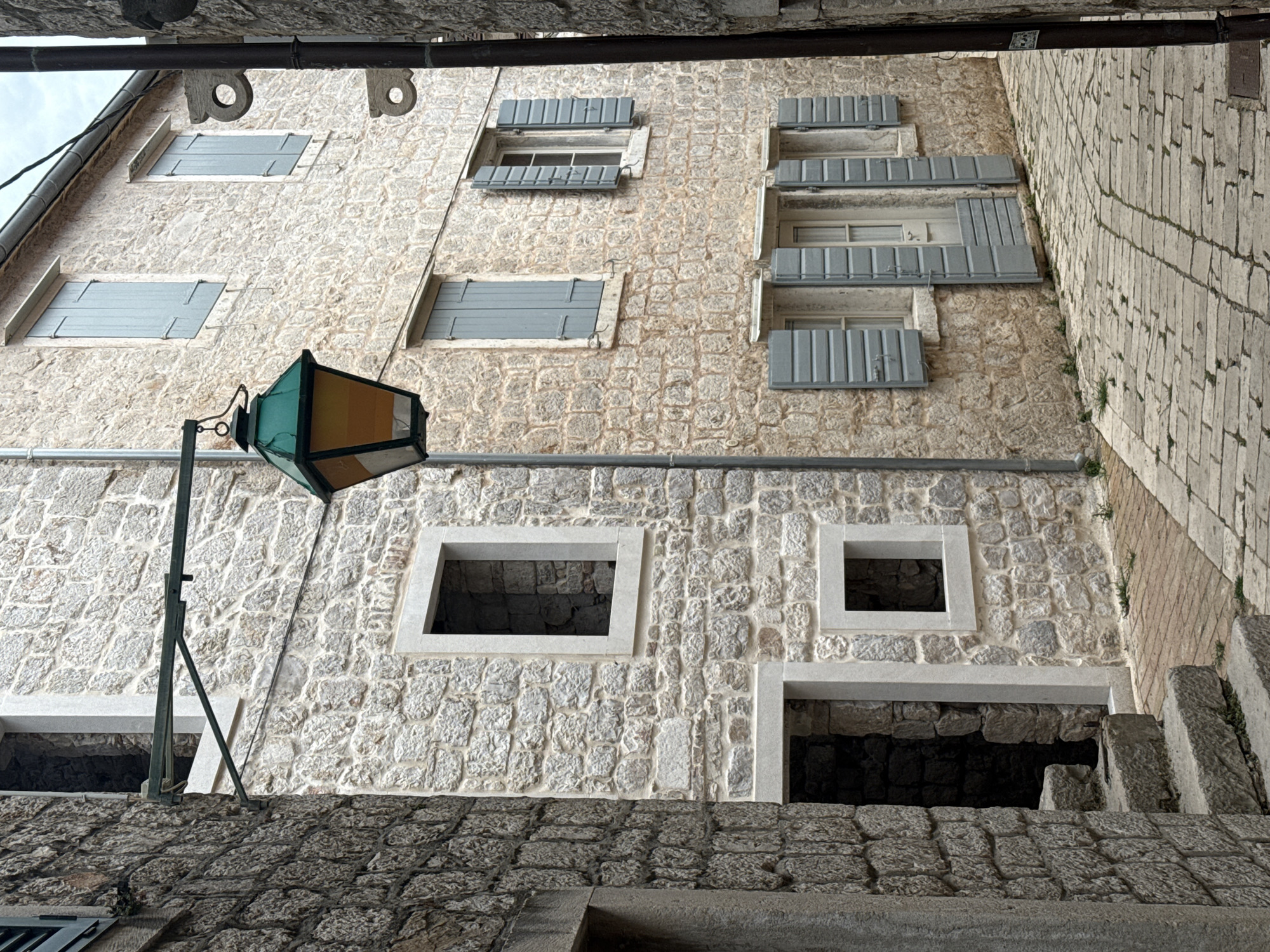 The image captures a narrow, cobblestone street in Grad Stari Grad, Croatia, flanked by traditional stone buildings. On the left, a building with rough-hewn stone walls features an open doorway with stone steps leading inside, and a small, square window opening above. To the right, a building with lighter-colored, more uniform stone blocks showcases several windows with pale blue shutters. A vintage lantern with a green hexagonal roof and amber glass panels hangs from an ornate metal bracket extending from the left building, casting a warm glow in the overcast lighting. The street is paved with interlocking stones, and a few sprigs of green grass sprout from the gaps between them. The architecture suggests a historic European setting, characterized by rustic stone construction and functional, aged design elements.