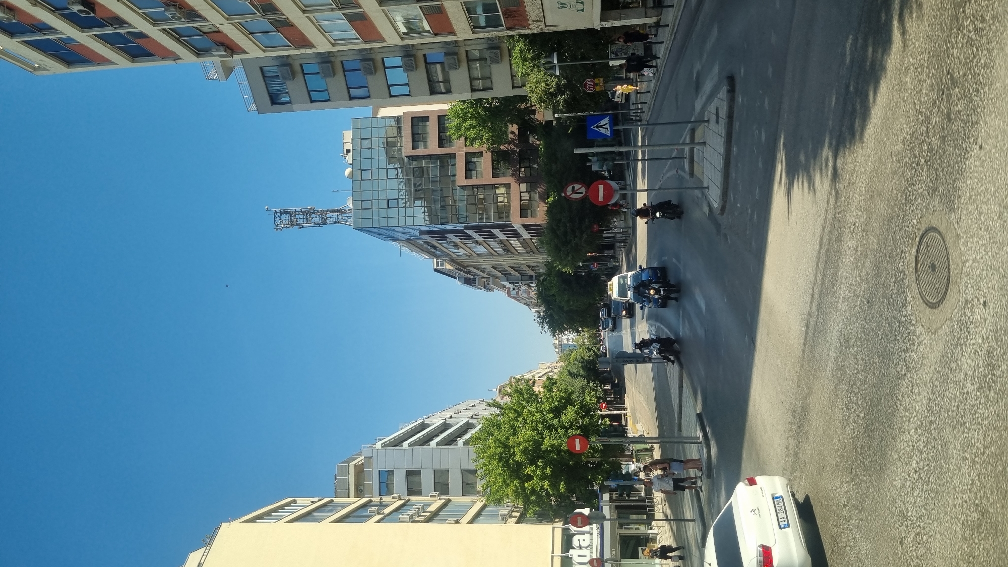 The image depicts a wide city street in Thessaloniki, Greece, under a clear blue sky. The street is lined with multi-story buildings, varying in architectural style, with some featuring modern glass facades and others displaying more traditional balconies. A prominent cellular tower is visible on top of a building in the distance.
The road is asphalt, with traffic lanes marked. Several vehicles are present, including a white car in the foreground with a license plate reading "AA 284 MA", a blue car, and several motorcycles. People are visible on the sidewalks, with a small group standing near the left side of the road. Traffic signs, including "stop" signs and directional signs, are placed along the street. Green trees are planted along the sidewalks, providing shade and contributing to the urban greenery. The scene appears to be captured during daylight, with shadows cast by buildings and trees indicating the time of day. The overall impression is a busy urban environment in Thessaloniki.