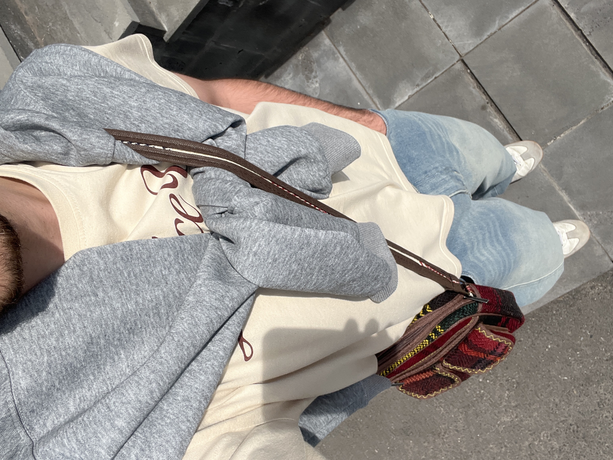 A person is standing on a Yerevan, Armenia sidewalk that has interlocking grey rectangular pavement slabs. The person is wearing light blue jeans, light grey shoes, a light tan shirt with brown lettering, and a light grey hooded sweatshirt. Across the person's body is a brown-strapped woven fabric bag. In the background is a black metal fence.