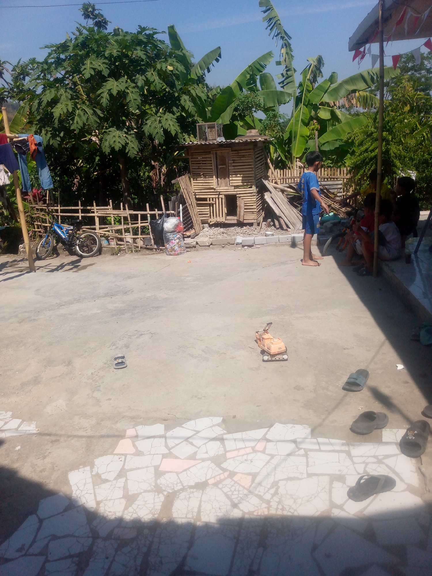 In Giriasih, Indonesia, a bright, sunny day is captured with a clear blue sky above lush green foliage. The foreground is a dusty, concrete ground scattered with broken white and pink tiles and several pairs of sandals, suggesting a casual, outdoor living space. A child's orange toy excavator sits on the concrete. In the middle ground, a blue bicycle is parked next to a woven bamboo fence. Behind the fence, laundry hangs on a bamboo pole. A small, wooden structure, possibly a coop or shed, is situated amidst the bamboo. Several children are visible: one boy in a blue t-shirt and shorts stands near the structure, while others sit or stand under a makeshift awning adorned with red and white triangular flags, their interactions suggesting a playful, communal atmosphere. The overall scene depicts a simple, rural setting.