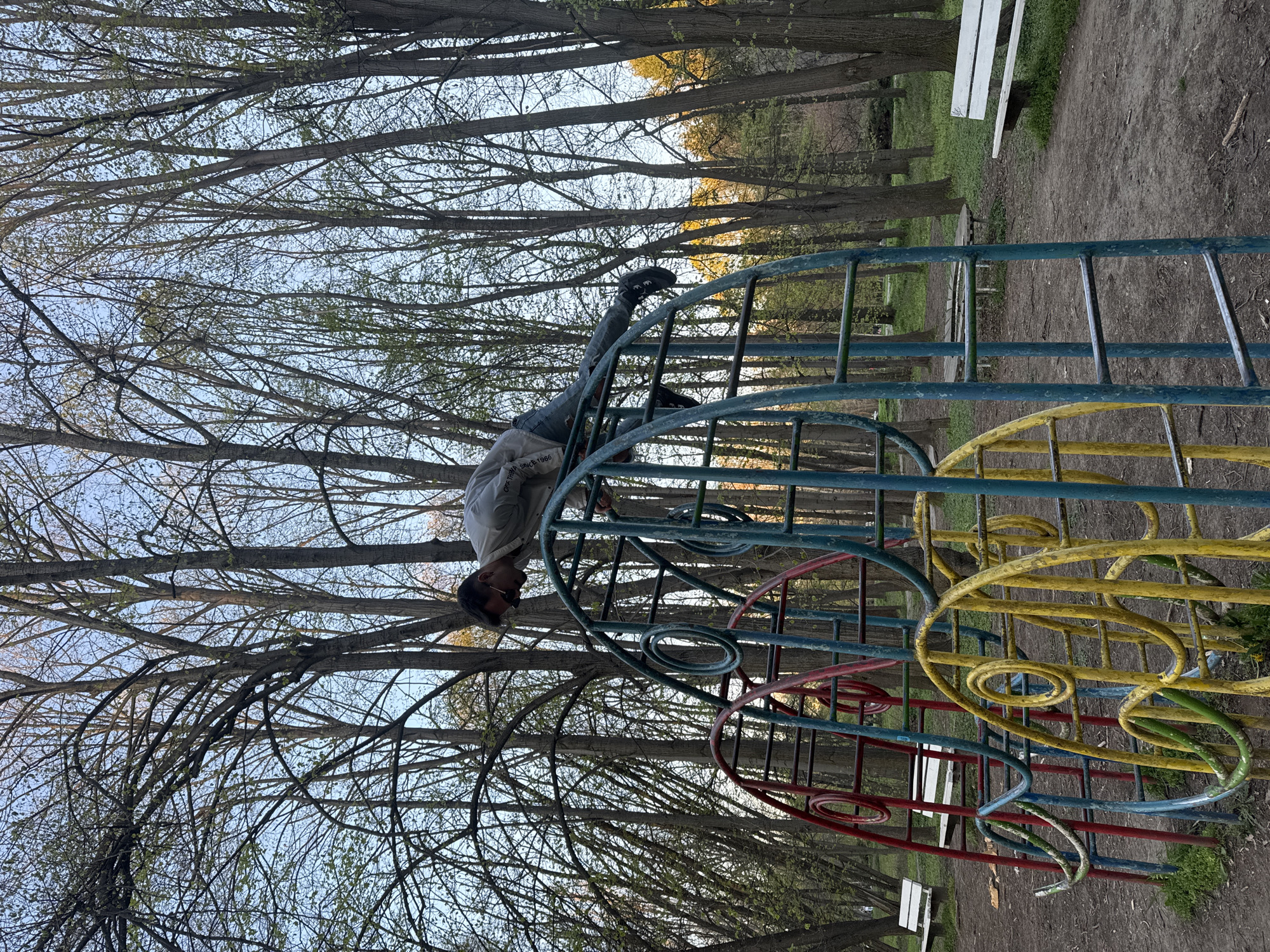 A person is hanging upside down on a colorful metal playground structure. The structure has blue, yellow, and red elements. The person is wearing a light-colored shirt, dark pants, and dark shoes with light-colored logos. They are gripping the top of the blue metal bars with both hands, their feet hooked on a higher rung. Behind the playground, there are numerous bare trees and a park environment. White benches are visible in the background. The image was likely taken in a park, possibly in Sofia, Bulgaria.