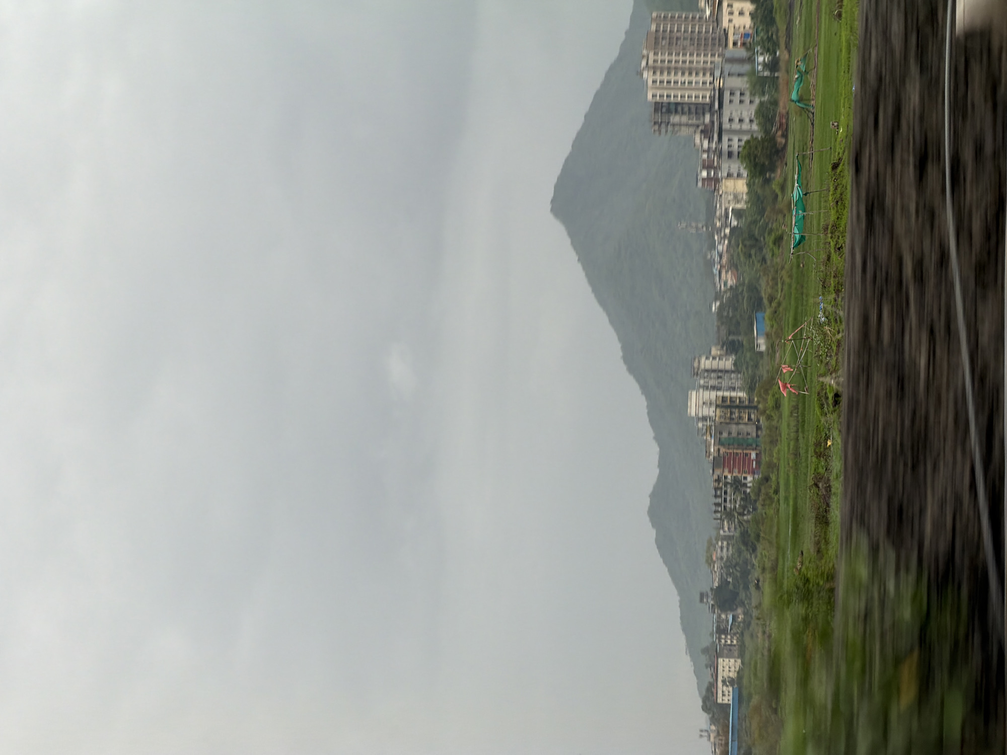The image captures a view of the city of Vasai-Virar, India, under a cloudy sky. A prominent, verdant mountain range forms the backdrop, its slopes softened by the overcast weather. In the midground, a cluster of buildings, ranging from residential complexes to smaller structures, is visible, nestled against the base of the hills. These buildings display a variety of architectural styles and colors. In the foreground, a lush green field stretches out, dotted with some agricultural elements like what appear to be small, temporary shelters covered with green tarpaulin, and what might be scaffolding or supports for some kind of structure. Two small, pink objects, possibly mannequins or decorative items, are positioned in the field, adding a touch of color to the otherwise natural and urban landscape. The foreground also shows a blurred section that suggests the image was taken from a moving vehicle, with a dark, textured element in the immediate foreground.