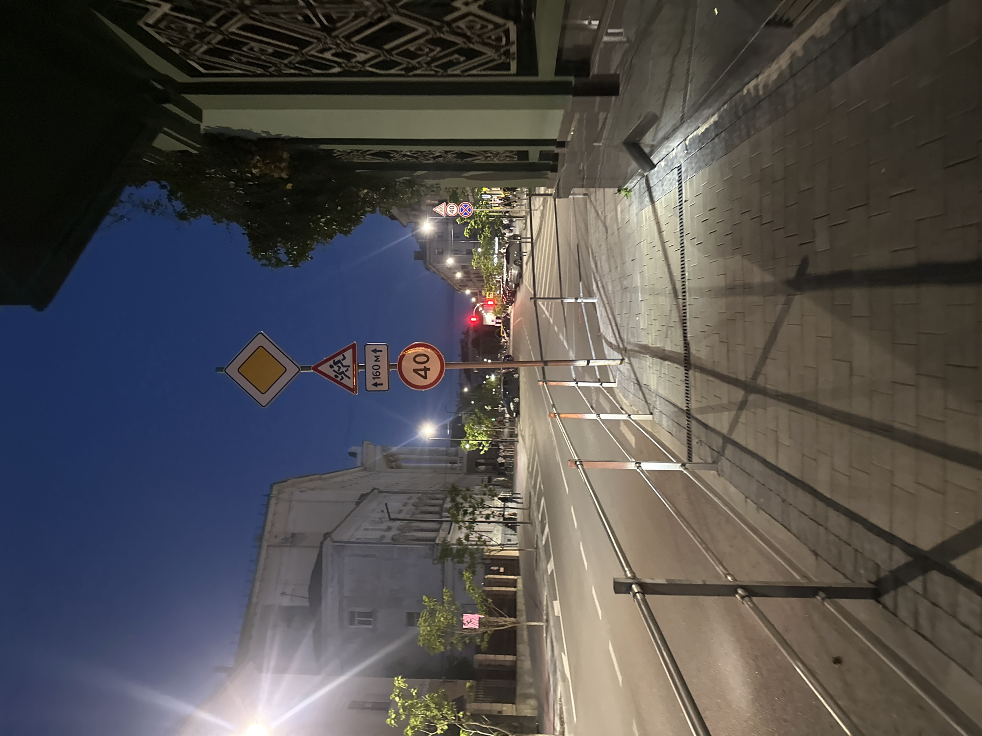 The image shows a street scene in Ternopil, Ukraine, at twilight or night. A wide, paved road runs horizontally across the frame, with parked cars and traffic lights visible in the distance. On the right side of the road, a paved sidewalk runs parallel to the street, bordered by a metal railing. Buildings line the left side of the street, and trees are planted along the sidewalk. Several traffic signs are visible, including a yield sign, a pedestrian crossing sign, and a speed limit sign displaying "40". The sky is dark blue, indicating a low light condition, and streetlights cast long shadows on the sidewalk.