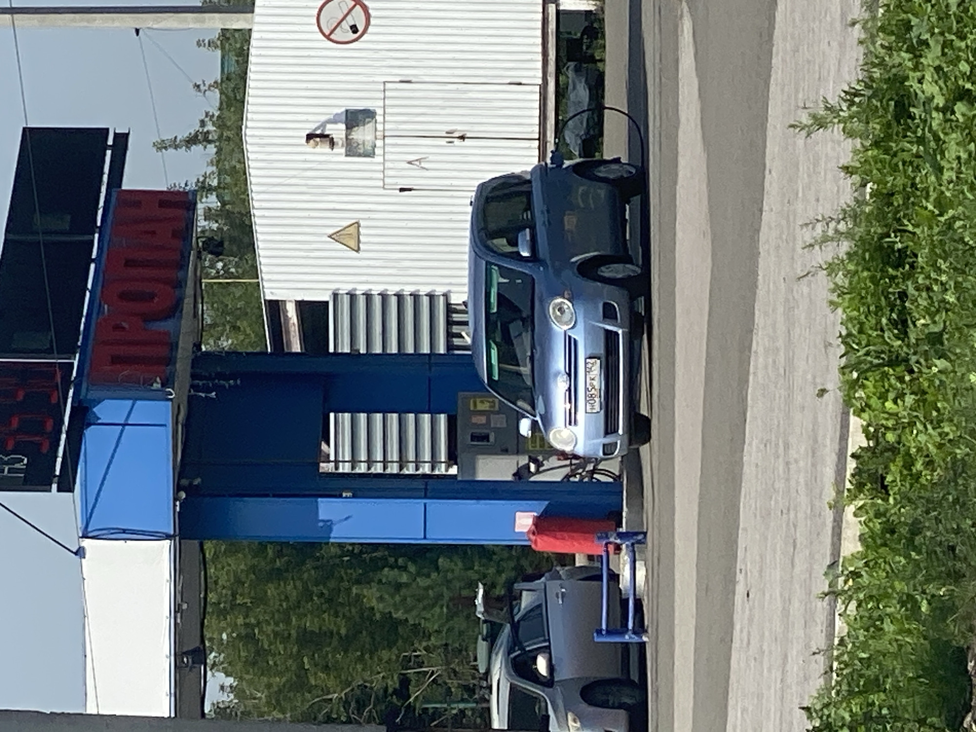 A blue minivan is parked at a gas station with a fuel hose connected to it. The minivan has a license plate that reads "H085PK 142". Next to it is a silver SUV with its driver's side door open. In the background, a white corrugated metal building stands next to a blue gas pump structure. A sign above the gas pump reads "ПРОПАН" (PROPANE). The scene appears to be a sunny day in Novokuznetsk, Russia, with trees visible in the background.