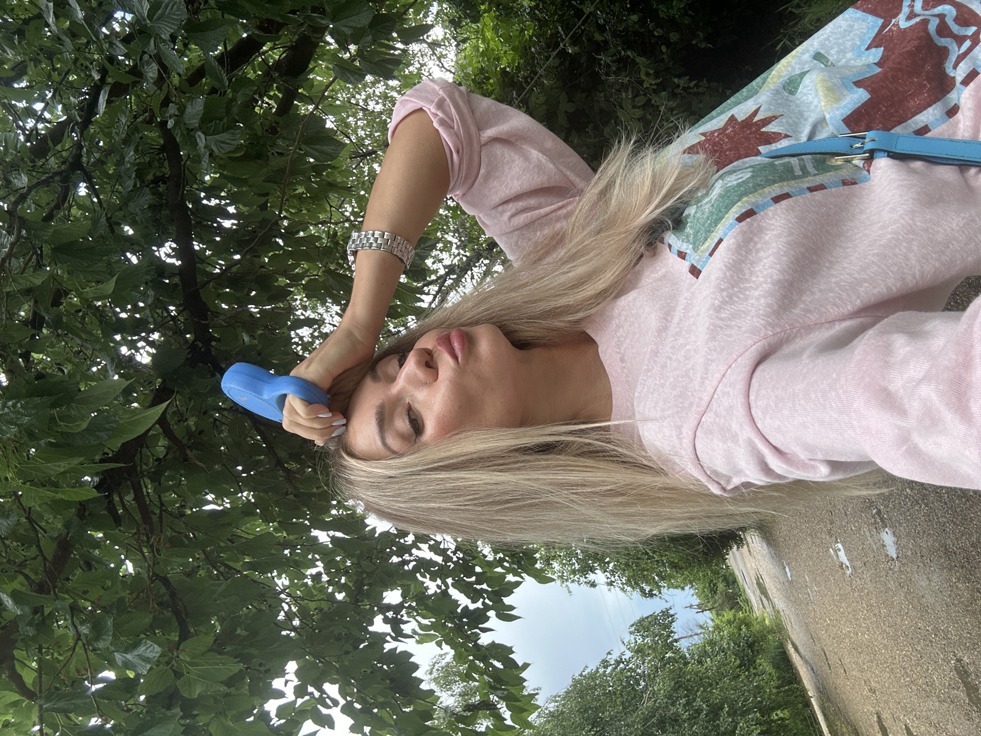An image captures a woman with long blonde hair standing outdoors near a gravel path with puddles, likely in Armavir, Russia. She is wearing a light pink long-sleeved shirt with a colorful graphic design and a silver watch. The woman is holding a blue object to her forehead and puckering her lips, as if posing for a selfie. Overcast skies and lush green foliage suggest it might be a cloudy day. The scene appears to be in a rural or semi-rural setting, with trees and vegetation bordering the path.