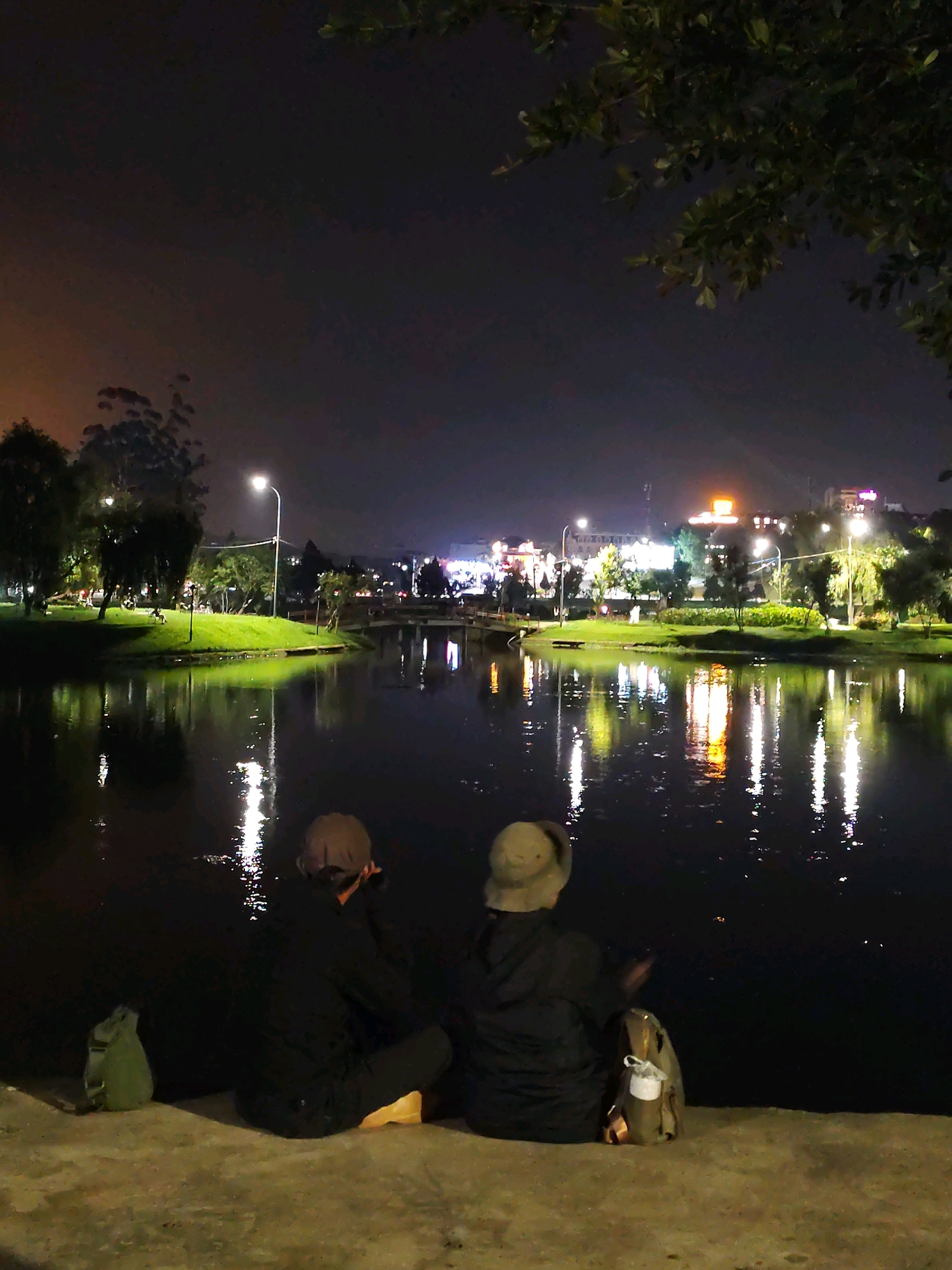 This image depicts Xuan Huong Lake in Da Lat, Vietnam, at night. The scene shows two individuals, seen from behind, sitting on the concrete embankment facing the lake. They are dressed in dark clothing and hats. One person has a light-colored hat, and the other a darker one. Beside them, to the left, is a green backpack, and to the right, a backpack with a white pouch attached. The lake's surface reflects the lights of the city and streetlights scattered across the opposite bank. The city skyline is visible in the background, illuminated by numerous lights from buildings and what appears to be a large, lit sign on a building in the center. Trees line the lake's edge on the far side, and a bridge is partially visible. The sky is dark, indicating nighttime.