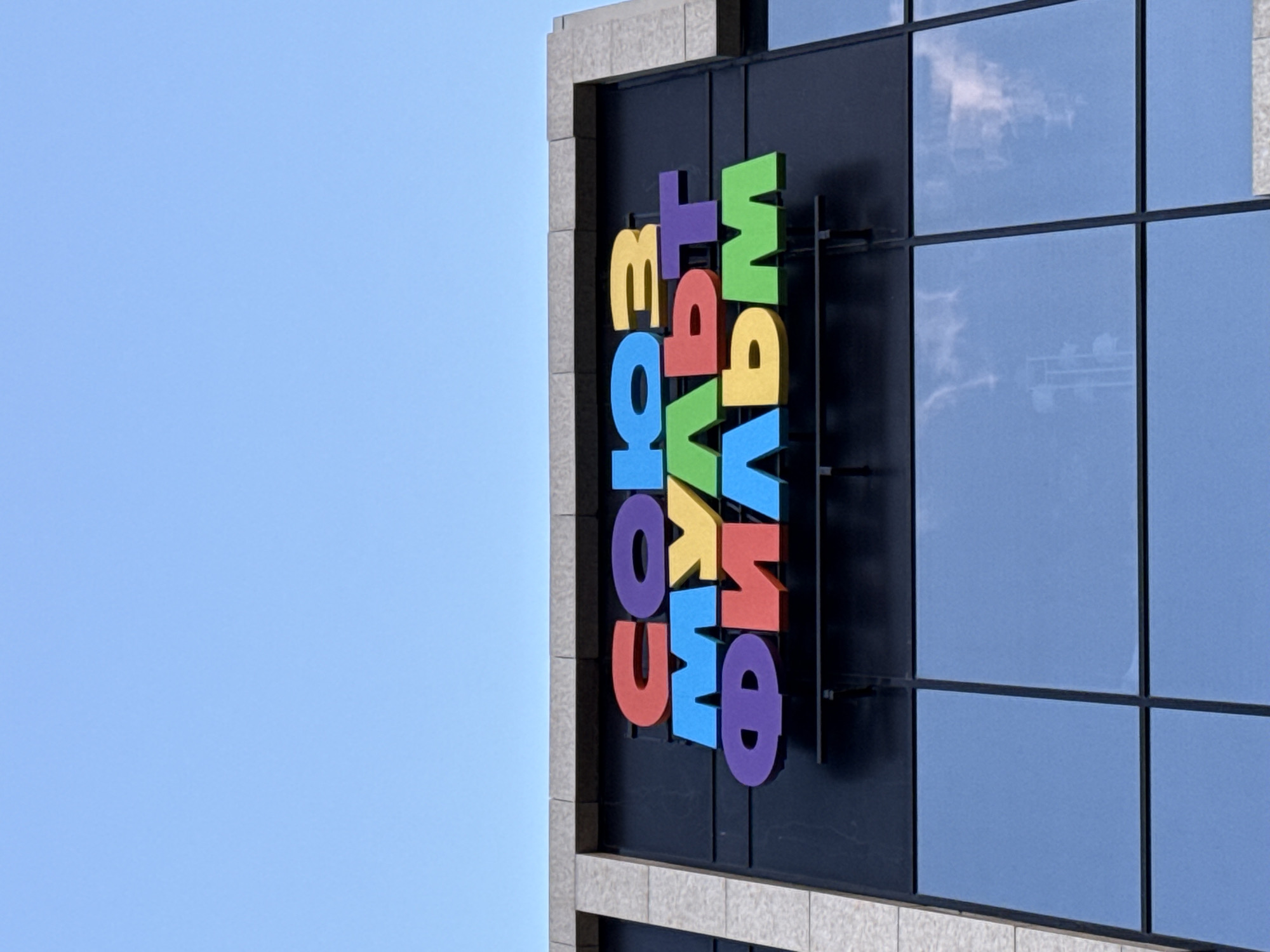 A clear, bright blue sky fills the left side of the frame. On the right, a modern building with a facade of dark glass panels and light grey, stone-like trim is visible. A large, colorful sign with three-dimensional Cyrillic letters is mounted vertically on the building's dark facade. Due to the original image's rotation, the building appears sideways and the sign's letters horizontally.
The sign spells out "СОЮЗ МУЛЬТ ФИЛЬМ" (Soyuzmultfilm), with each letter in a different vibrant color: "С" is blue, "О" is red, "Ю" is orange, "З" is yellow, "М" is purple, "У" is green, "Л" is red, "Ь" is blue, "Т" is yellow, "Ф" is red, "И" is blue, "Л" is purple, and "ЬМ" is green. Some letters exhibit slight shadows, indicating they project from the building surface.
The building's glass windows reflect parts of the sky, showing soft white clouds and what appears to be a distant structure or antenna array. The day is sunny and clear, with no visible activity or people. The architecture suggests an urban or corporate setting.