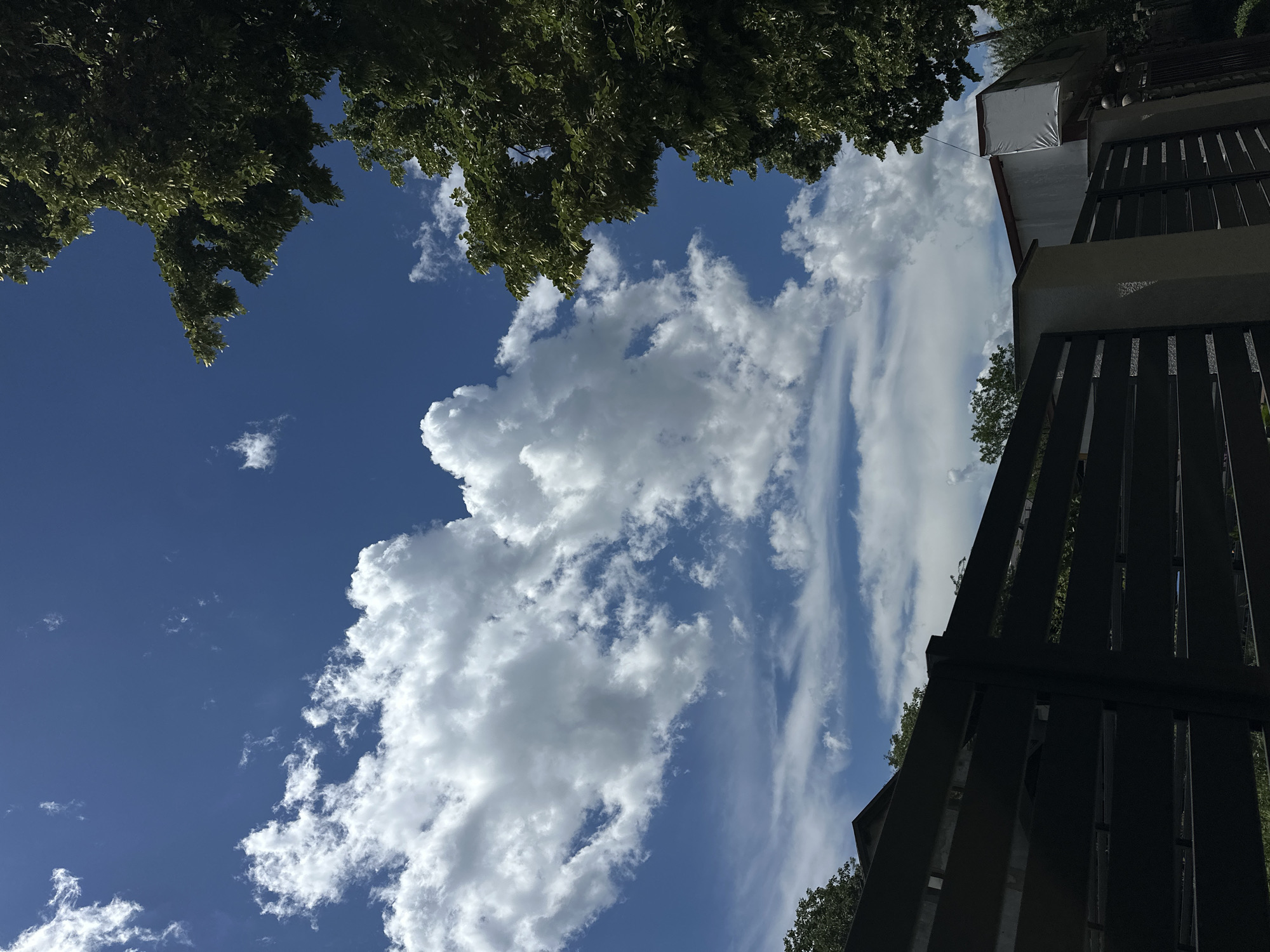 The image shows a view of the sky from a residential area in Strășeni, Moldova. The sky is a deep blue with scattered white cumulus and cirrus clouds. In the foreground, there is a dark horizontal fence made of metal slats, partially obscuring the view of a building with a white exterior and a red roof. To the right, a large tree with green foliage partially frames the scene. The overall impression is one of a sunny day in a suburban or rural setting.