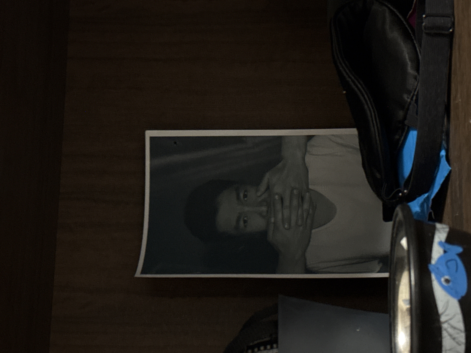 The image shows a black and white photograph of a person covering their mouth with their hands. The photograph is propped against a dark wooden surface, possibly a shelf or drawer. A black bag with blue accents is partially visible next to the photograph. A black can with a blue fish sticker is also visible. The location of the scene is not evident from the image. The city of Khimki, Russia cannot be identified as a location.