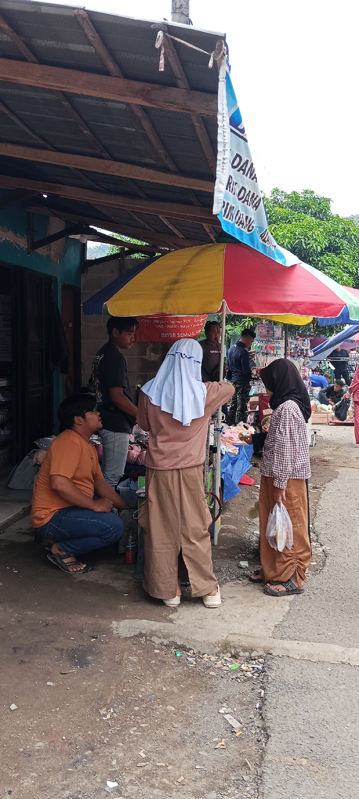 The scene takes place in a street market in Kecamatan Kutawaringin, Indonesia, located at coordinates -6.973 latitude and 107.52 longitude. A large colorful umbrella provides shade for a stall with various items for sale. Four adults are present. One man, wearing an orange shirt and blue jeans, is crouched on the ground, seemingly talking to another man wearing a black shirt and jeans. A woman, wearing a brown dress and white headscarf, stands near the back of the stall, facing the other man. A woman wearing a checkered top and a brown headscarf is standing near the stall, holding a plastic bag. The stall is set up along a concrete sidewalk with a dirt and gravel road to the right. A sign with the words "DANA" and "RIK DANA" is visible on the building to the left. Several small items, including cigarette butts, are scattered on the ground around the stall.