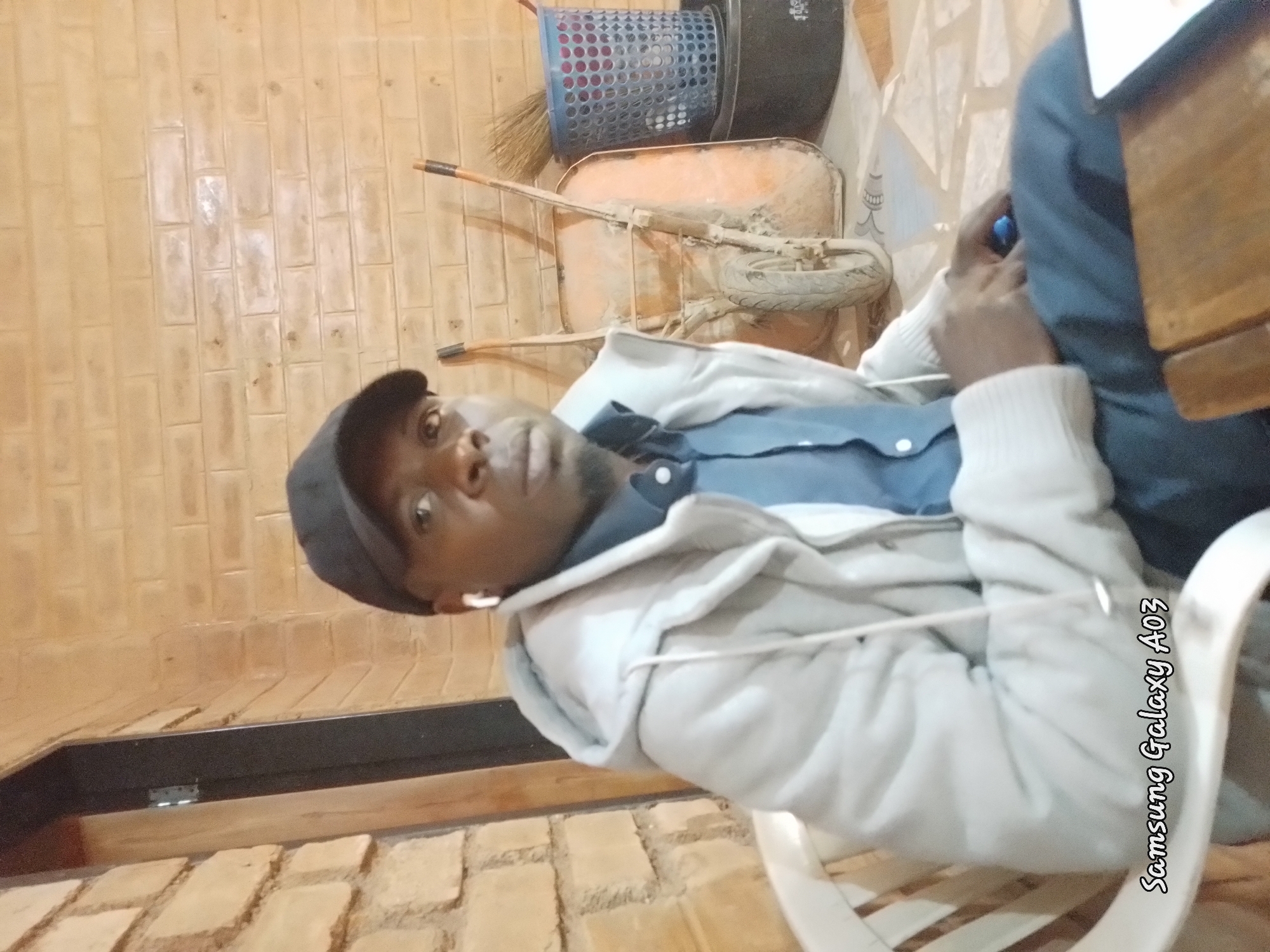 A medium shot captures a Black male wearing a black baseball cap, a light gray hooded jacket over a blue collared shirt, and blue pants. He is seated in a white chair, looking towards the viewer with a neutral expression. White earbuds are visible in his ears. He holds a small blue object in his right hand. In the background, a brick wall forms the backdrop, with a wheelbarrow and a blue basket containing a broom to the right of the subject. The lighting is warm and appears to be from artificial sources. The scene is set outdoors, possibly in a yard or patio area. The bottom left corner of the image displays "Samsung Galaxy A03" text.