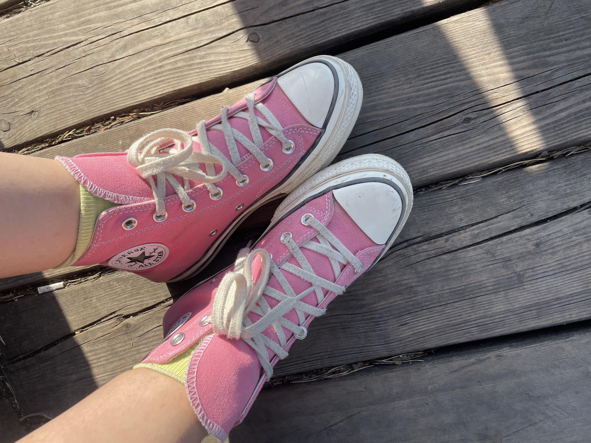 A pair of pink high-top Converse sneakers are positioned on a wooden surface. The left sneaker's side faces the camera, revealing the Converse logo, while the right sneaker is angled slightly away. Both sneakers are laced with white laces tied in a casual knot. The wooden surface appears to be made of planks, with varying tones of brown and grey. Shadows are cast across the wood, suggesting an outdoor setting. The wearers legs and socks are visible. The style of the shoes and surface do not provide definitive evidence to place the location in Moscow.
