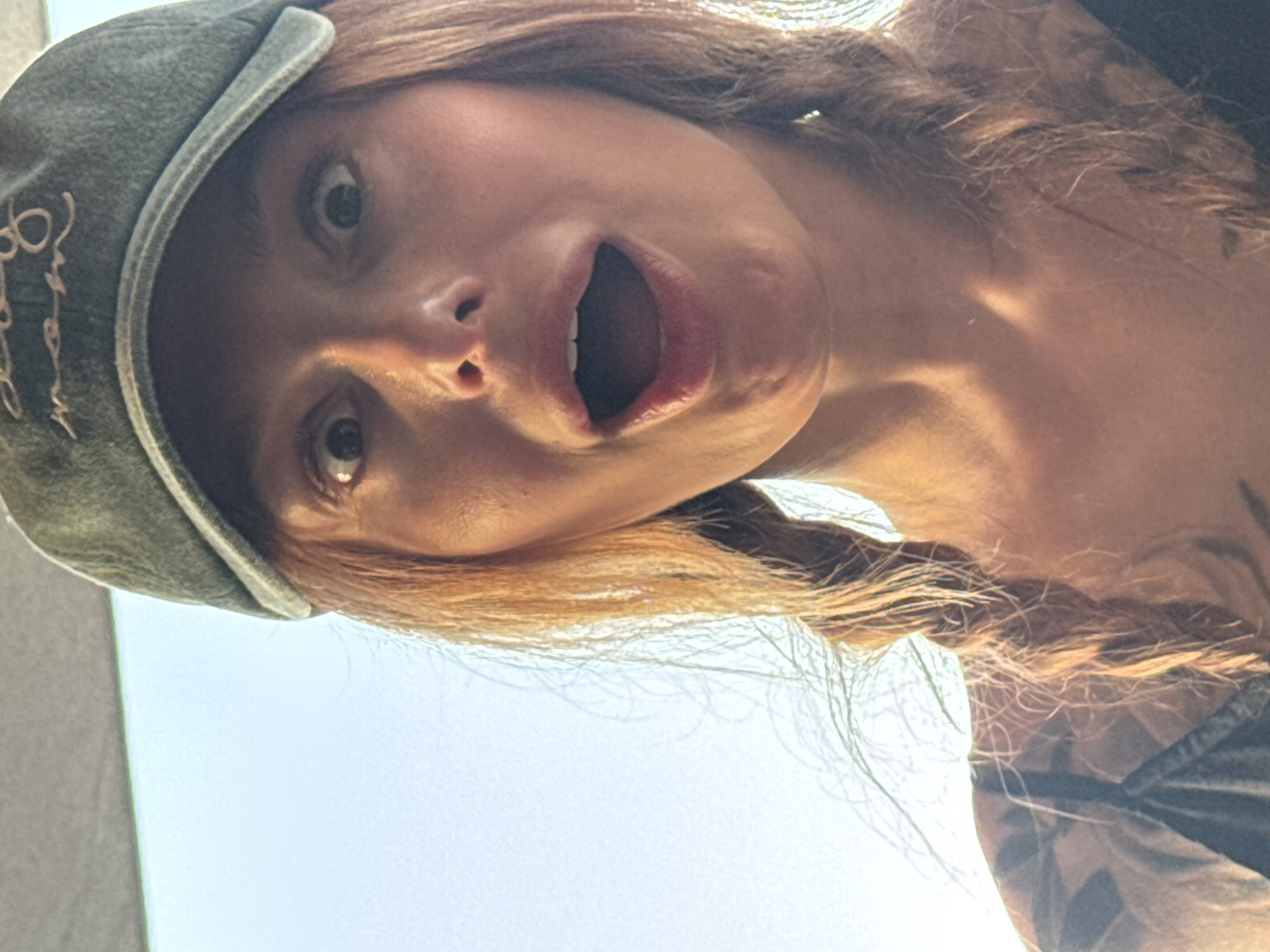 A woman with light brown hair and a shocked expression is looking directly down at the camera. She has her mouth open wide and her eyes are wide. She is wearing a green cap with embroidered text on the left side of the brim. Her hair is in braids. Her upper chest and a portion of her left shoulder are visible, revealing a tattoo. The background is primarily bright sky. The location is Bogišići, Montenegro.