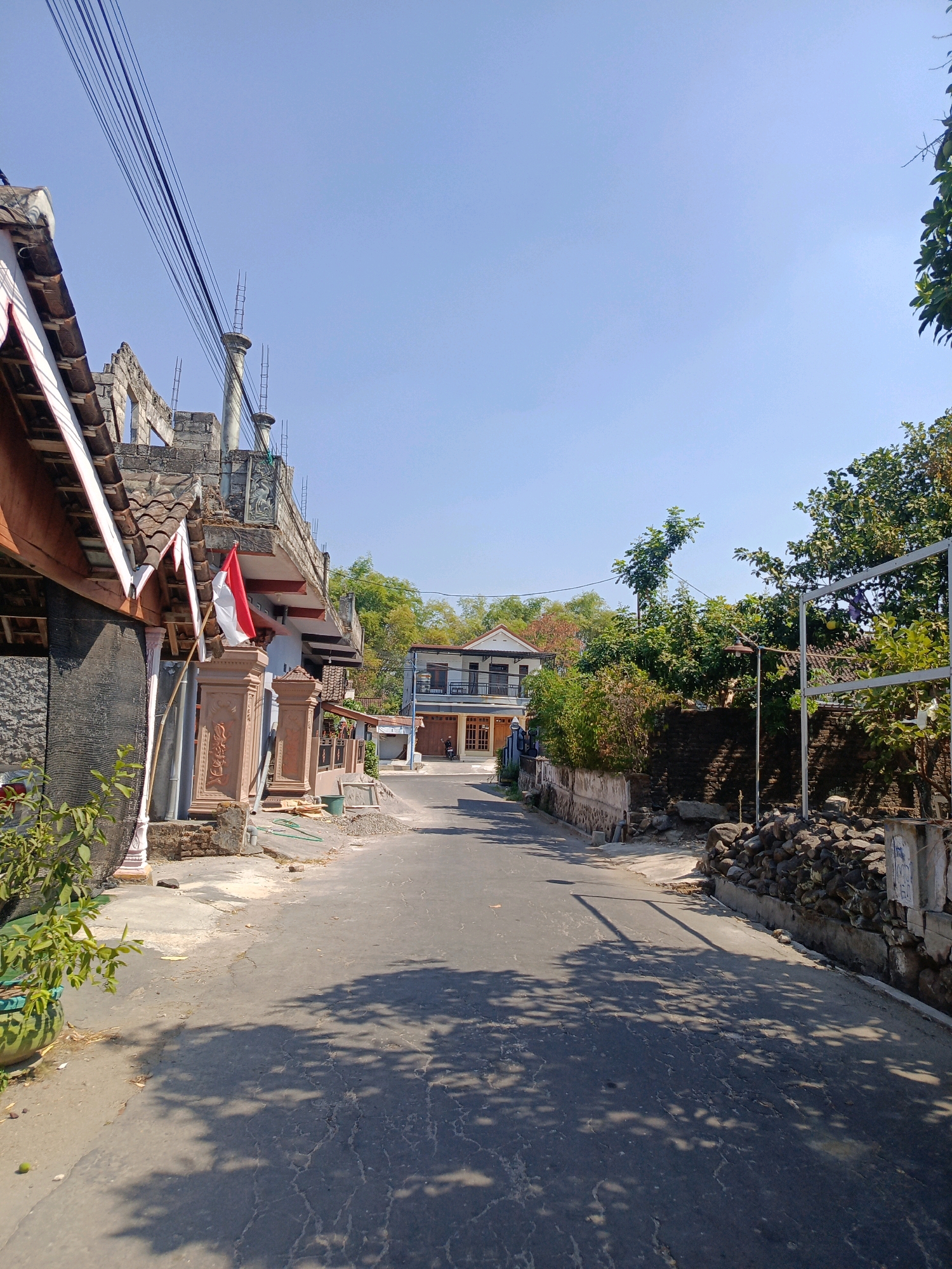 The image depicts a street scene in Tambakrejo, Indonesia, under a clear blue sky. The road surface is asphalt, showing signs of wear and tear with visible cracks and patches, and is shaded by dappled tree shadows. On the left side of the street, a building under construction is visible, with exposed concrete pillars and rebar. A small Indonesian flag, red and white, is prominently displayed on a decorative pillar in front of this building. Adjacent to the construction site are other residential buildings with tiled roofs and various architectural features.
The right side of the street is lined with a low stone wall, behind which vegetation and trees grow. Piles of rocks are also present along this side. Further down the street, more houses and buildings can be seen, suggesting a residential area. A motorcycle is parked in front of one of the houses in the distance. The overall environment appears to be a typical street in a moderately developed Indonesian town, with a mix of construction, residential properties, and natural greenery. The time of day appears to be daytime, given the strong sunlight and shadows. There are no people visible in the image.