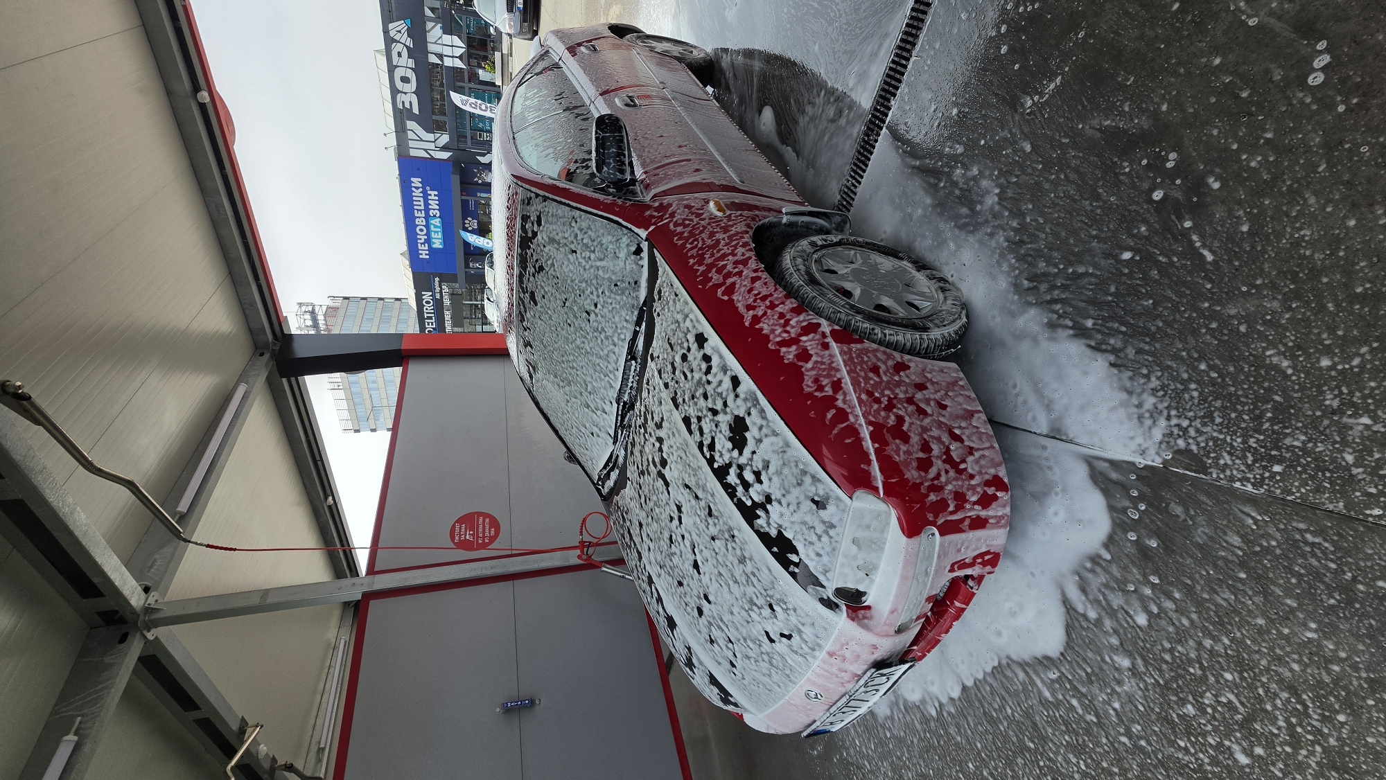 The image depicts a red car being washed at a self-service car wash. The car is covered in white soap foam. A high-pressure water hose is directed at the car, spraying it. The car wash is located in an urban area, identifiable by the presence of modern buildings and commercial signage in the background. Specifically, the Bulgarian sign "НЕЧОВЕШКИ МЕГАЗИН" is seen; it can be loosely translated to "Inhuman Megastore", as well as another commercial sign "Zora". The location is Varna, Bulgaria, based on the car's license plate. The ground is wet and covered in soap suds. The structure of the car wash is visible, including the metal frame and roof.