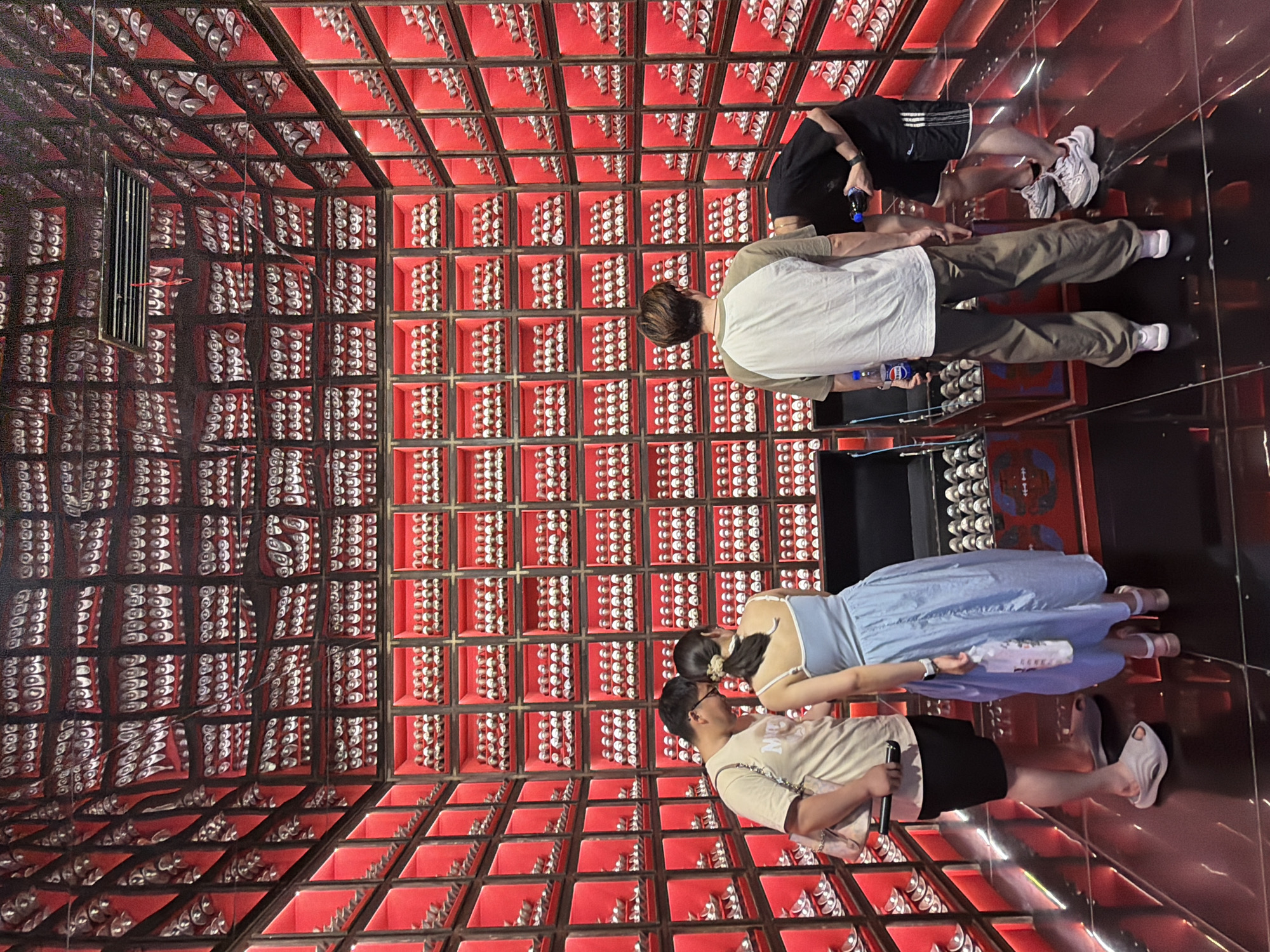 In Yuzhong District, China, a man and a woman stand facing each other in front of a wall lined with numerous red shelves filled with small, uniform items. The man wears a light-colored t-shirt and dark shorts, while the woman is in a light blue dress. To their right, two other men are positioned near large, open containers also filled with the same small items. One of these men is wearing a grey t-shirt and baggy trousers, holding a bottle, and the other is in a black t-shirt and athletic shorts, bending down and also holding a bottle. The environment is characterized by the dominant red shelving system, the reflective black floor, and the overall symmetrical arrangement of the items on display.