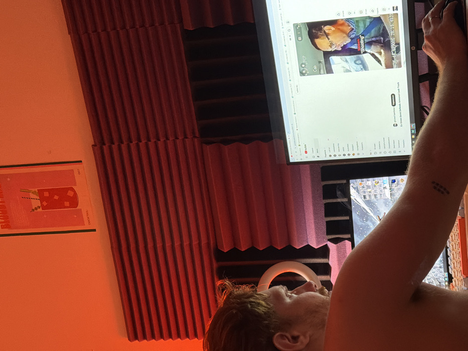 A person with ginger hair is seen from a low angle, facing a computer monitor. The screen displays a YouTube video featuring a man in glasses. A tattoo is visible on the person's arm. The background consists of soundproofing foam in alternating shades of red and dark gray. A poster with a cocktail image hangs on the wall beside the soundproofing. A ring light is situated near the person’s head. The overall lighting is warm, casting a reddish hue.