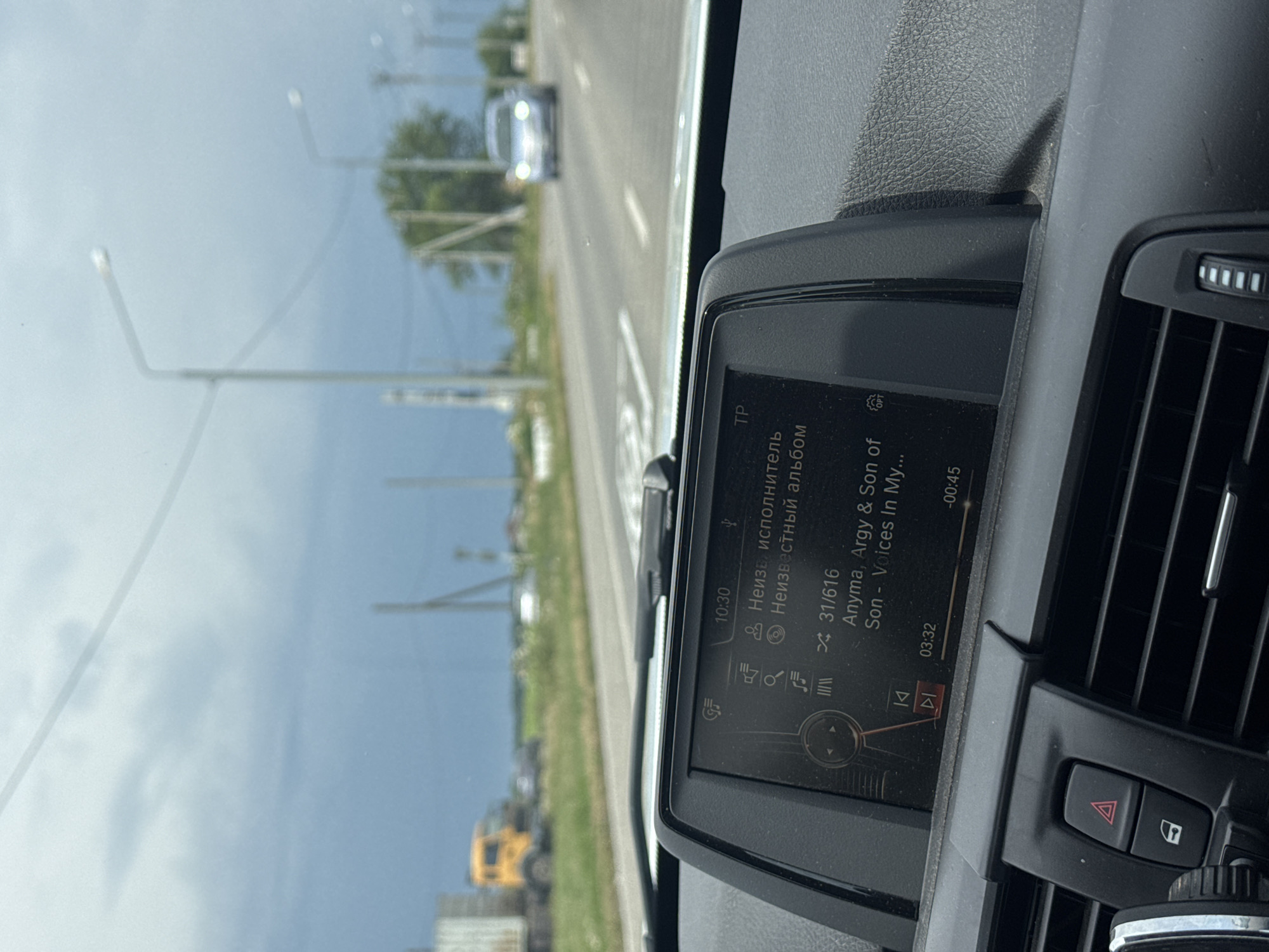 The image shows the interior of a car dashboard, with a digital display showing music information. The screen reads "10:30" at the top and displays song details including "Anymа, Argy & Son" as the artist and "Son - Voices In My..." as the song title. The track number is "31/616". Below the song title, the current playback time is "03:32" and the total track duration is "-00:45". The car is on a road, with a cloudy sky visible through the windshield. A yellow truck and a silver car are visible in the distance on the road. The image is taken from the perspective of the driver or a passenger in the front seat. The car's hazard lights button and a car door lock button are visible below the display. The overall environment appears to be outdoors, with a road and trees in the background.