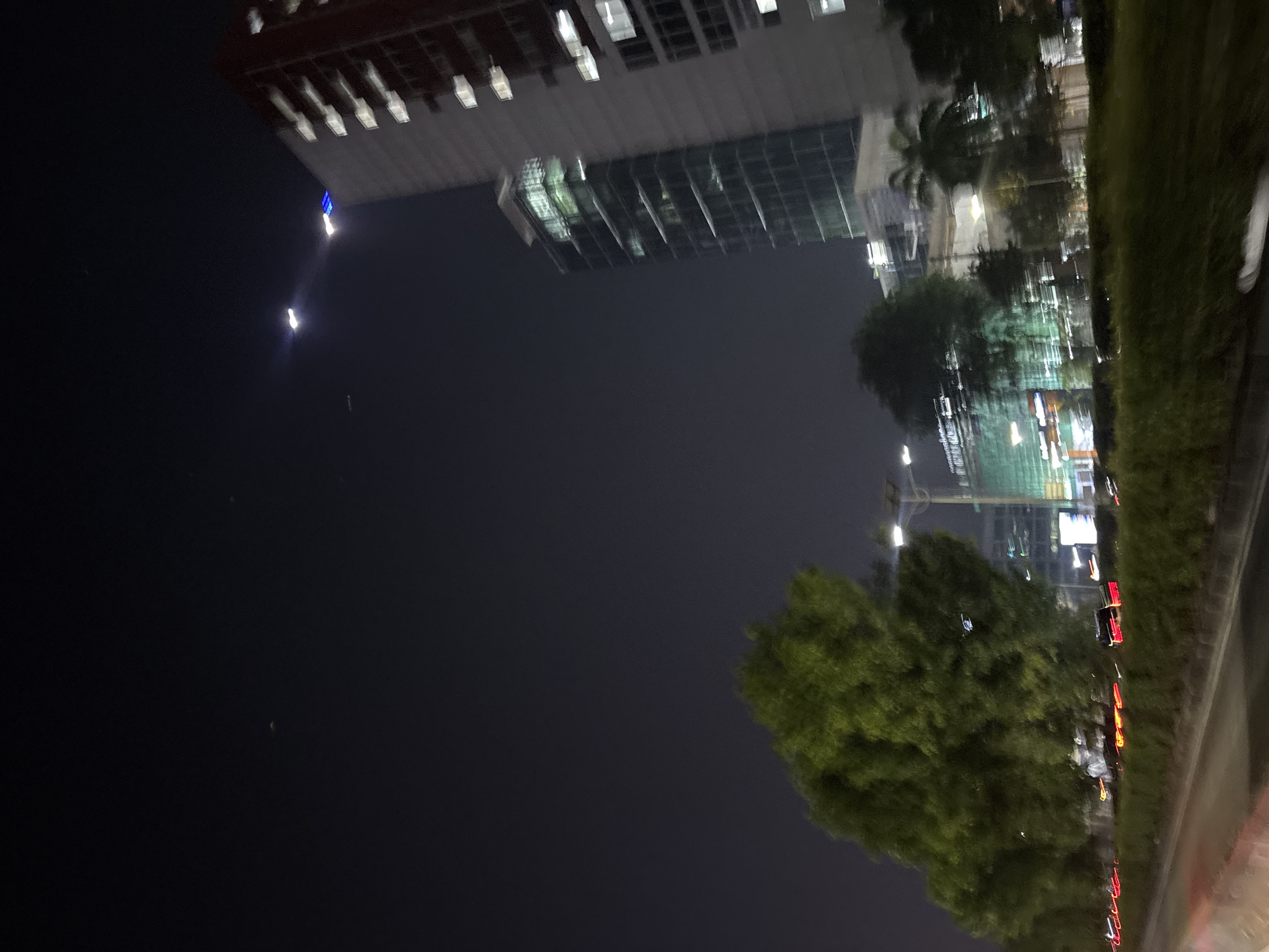 The image, taken at night in West Jakarta, Indonesia, depicts a street scene with a prominent modern building on the left. The building features numerous windows, some illuminated with a blue light at the top. Below this, a section of the building has a glass facade reflecting the lights of the city. A large, leafy tree occupies the foreground on the right side of the image, obscuring some of the background details. Further to the right, a grassy embankment runs alongside a road where red taillights of vehicles are visible, suggesting traffic. The sky is dark, indicating it is nighttime. The overall impression is of an urban environment at night, with a mix of commercial architecture and natural elements. There is no visible text in the image.