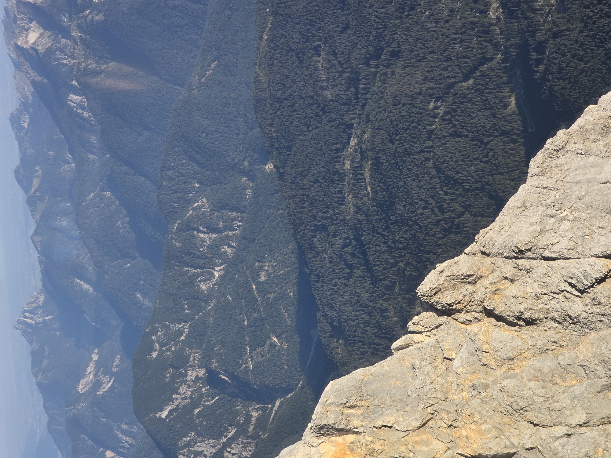 The image presents a panoramic view of a mountainous landscape, likely in the Bavarian Alps near Garmisch-Partenkirchen, Germany. The foreground features a sharply angled, sunlit rock face, exhibiting textured surfaces and warm, earthy tones. Beyond this immediate rocky prominence, the midground and background are dominated by a vast expanse of densely forested mountains. The trees, predominantly dark green, blanket the slopes, creating a textured carpet that recedes into the distance. Faint, winding paths or roads are subtly visible within the forest, suggesting human presence or access. The furthest mountains are softened by atmospheric haze, appearing as layered silhouettes of blue and gray, with their peaks gradually fading into a light blue sky. The scene is devoid of people or specific urban structures, focusing entirely on the natural grandeur of the alpine environment.