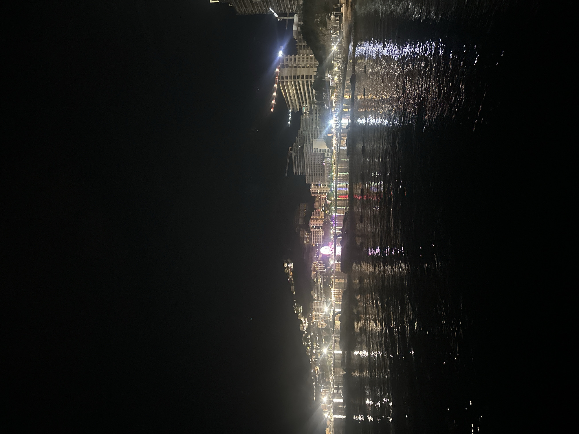 The image depicts a nighttime view of the city of Budva, Montenegro, observed from the sea or a coastal vantage point. The dark sky occupies the upper half of the frame. Below, the city's illuminated skyline stretches across the midground, rising from the shoreline up a gradual hillside. Numerous multi-story buildings, appearing as residential or commercial structures, are densely packed and heavily lit, their lights casting a warm glow. Towards the left-center of the cityscape, a large, brightly illuminated Ferris wheel stands out with prominent colorful lights, including purple. Further to the right, several tall construction cranes, with their booms and structures illuminated, indicate ongoing development along the coastline. The shoreline is defined by a continuous band of artificial lights, suggesting a lively promenade or beachfront establishments. The foreground is dominated by a dark body of water, likely the Adriatic Sea or Budva Bay. The water's surface reflects the city lights as elongated, shimmering streaks of white, yellow, and various other colors, including the purple hues from the Ferris wheel. Several indistinct, dark shapes consistent with boats or pontoons are partially visible on the water near the illuminated shore. No discernible human figures are present in the image.