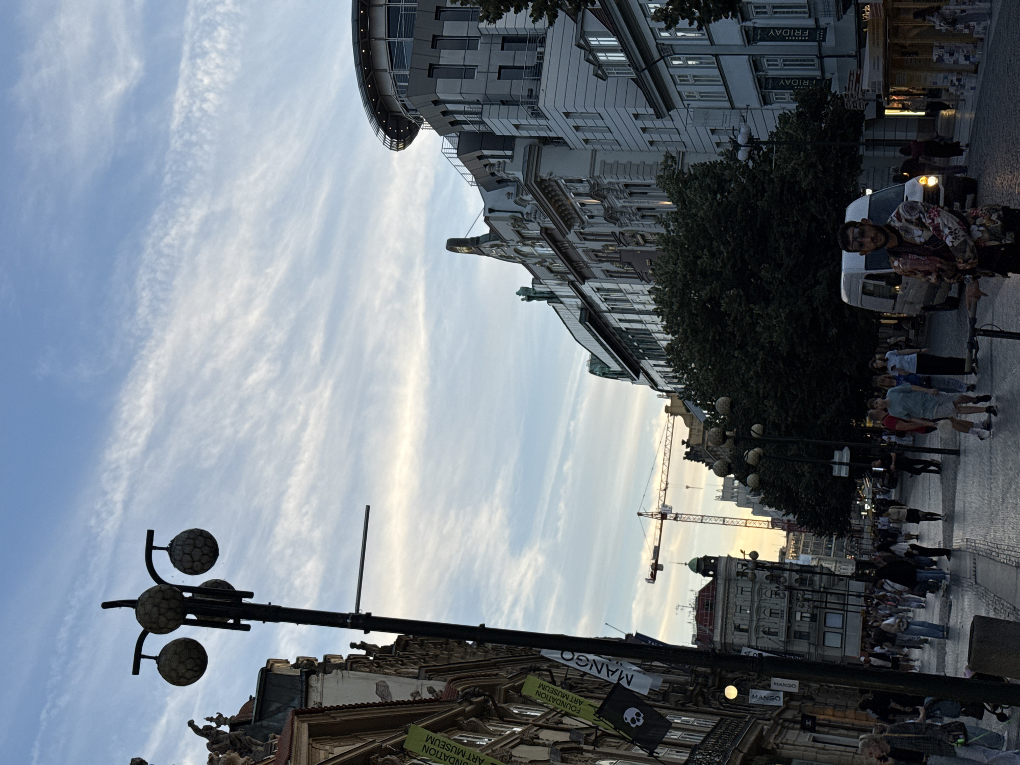 The image captures a street scene in Prague, Czechia, likely during the late afternoon or early evening. The sky is a soft blue with wispy white clouds. In the foreground, a dark lamppost with three spherical lights stands on the left side. To the left of the lamppost, a building facade is visible, adorned with banners. One banner features the text "FOUNDATION ART MUSEUM" and a skull and crossbones motif. Another banner reads "MANGO".
The street is paved with cobblestones and is populated by numerous pedestrians walking in both directions. A white van is parked on the right side of the street, and a man wearing a patterned shirt and sunglasses stands in front of it. Further down the street, more buildings line both sides, with trees interspersed. A large construction crane is visible in the distance. The architecture suggests an urban European setting, consistent with Prague's historic center.